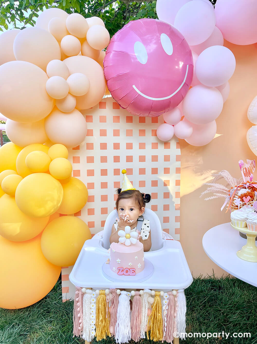 Momo Party One Groovy Baby First Birthday Party, with a birthday girl wearing a Retro Daisy Party Hat, sitting in the hair chair, eating her smash cake with a check patterned wooden Backdrop board, 30" Rosy Smiley face foil balloon by Tuftex Balloons, latex balloon decorations in peach, pink, Goldenrod colors. This decoration is such a stand-out backdrop and photo booth for a Groovy birthday party, baby showers, and almost any other joyful event