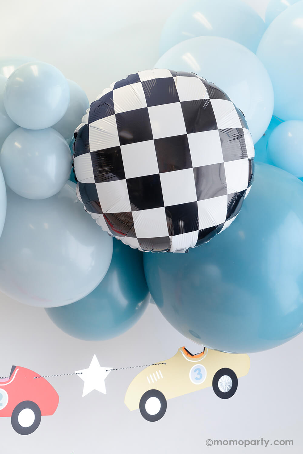 A modern race car themed party decoration by Momo Party featuring a blue balloon garland adorned with checkered foil balloons, underneath hung a vintage race car party garland in pastel colors by Meri Meri. Makes this a perfect party decoration for kid's "Two Fast" second birthday party or a "Fast One" first birthday party.