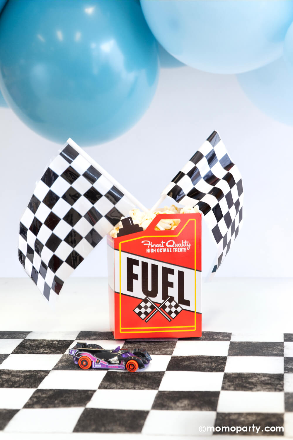 A fuel-treat-box topped with race car checkered flags from Momo Party with popcorn in side. It's setting on a checkered paper table runner with blue balloons hung on top - making this a perfect snack ideas for kid's race car themed birthday or a Hot Whees themed bash.
