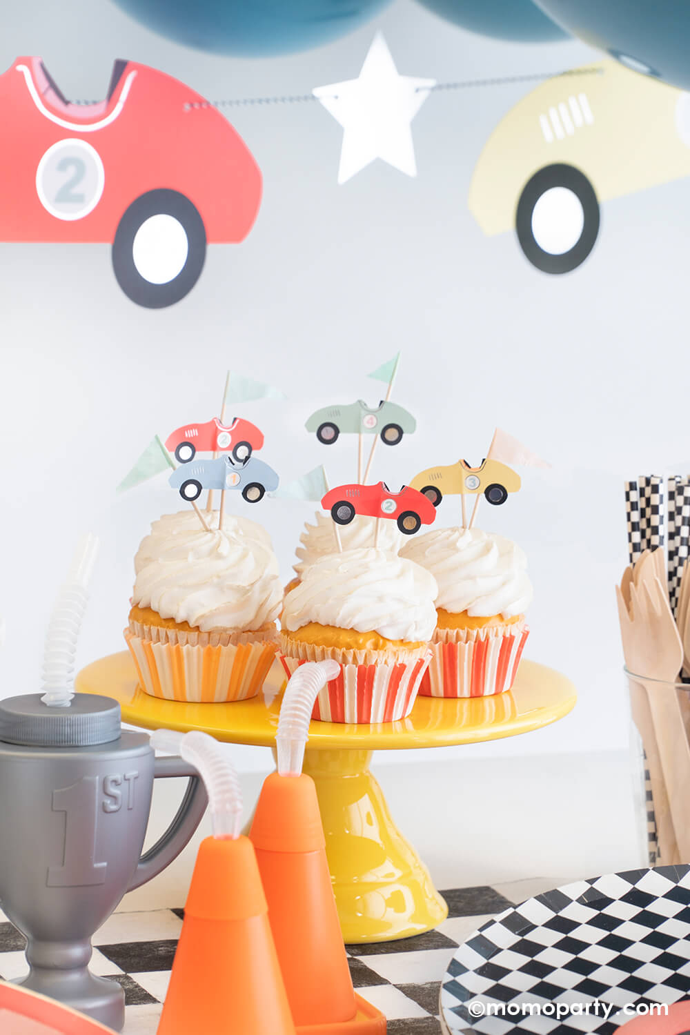 A festive kid's race car party set up featuring some cupcakes decorated with Momo Party's race car cupcake kit that comes with vintage race car toppers and flag toppers in pastel colors, along with orange and red striped cupcake wrappers. Around the cupcakes are some cone shaped and trophy shaped tumbler sippers and round checkered plates, table runner and party straws. Above the table is Meri Meri's race car party garland with some party balloons.