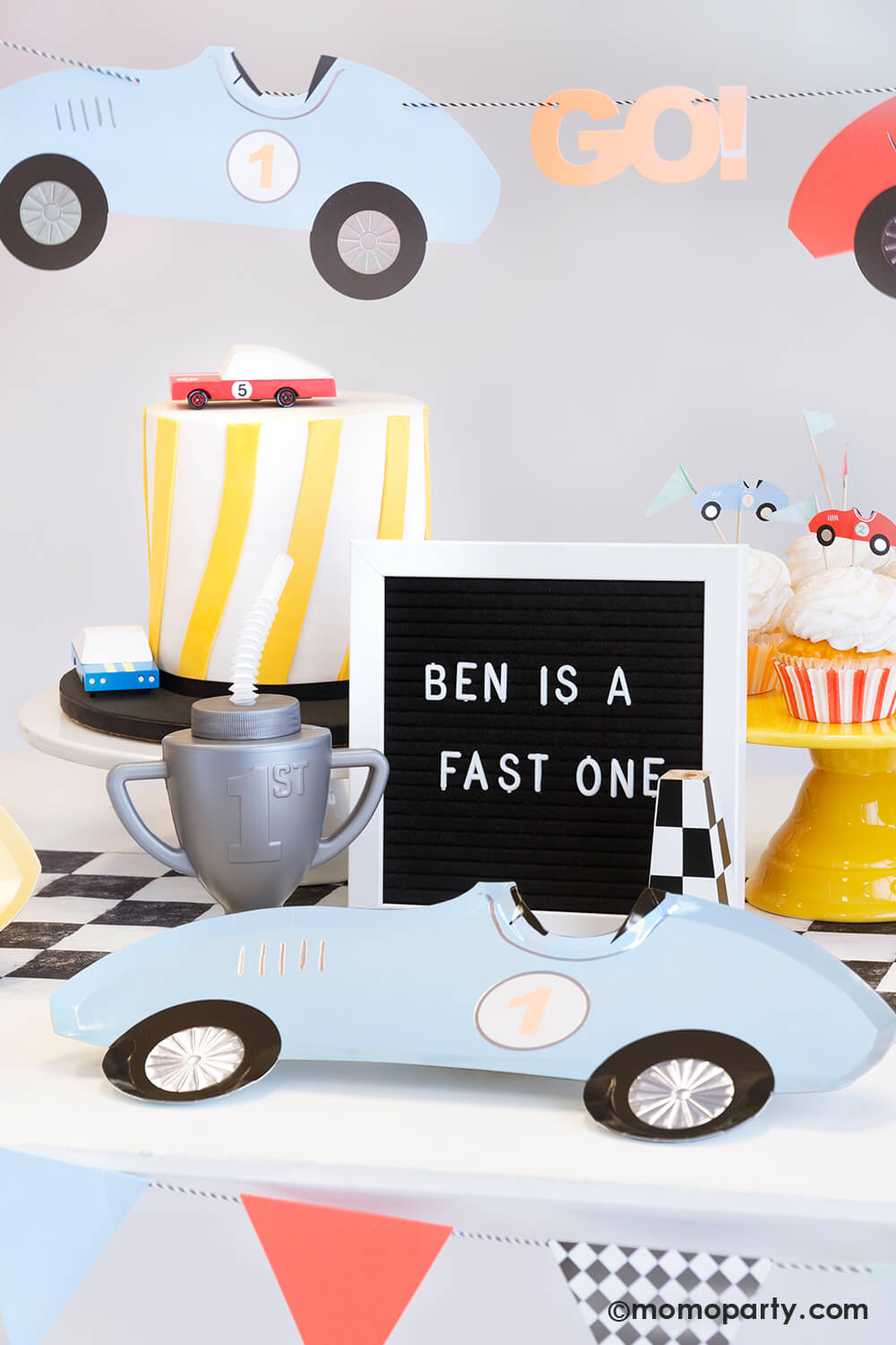 A "Fast One" race car themed first birthday party table featuring Momo Party's race car shaped paper plates, a trophy shaped sipper on a checkered paper table runner, along with a modern cake adorned with wooden toy race cars and some cupcakes decorated with Meri Meri's race car toppers. Above the table hung a vintage race car party garland in soft pastel colors. Along with a letter board spelling "Ben is a fast one" , it makes a perfect inspiration for a baby boy's first birthday celebration.