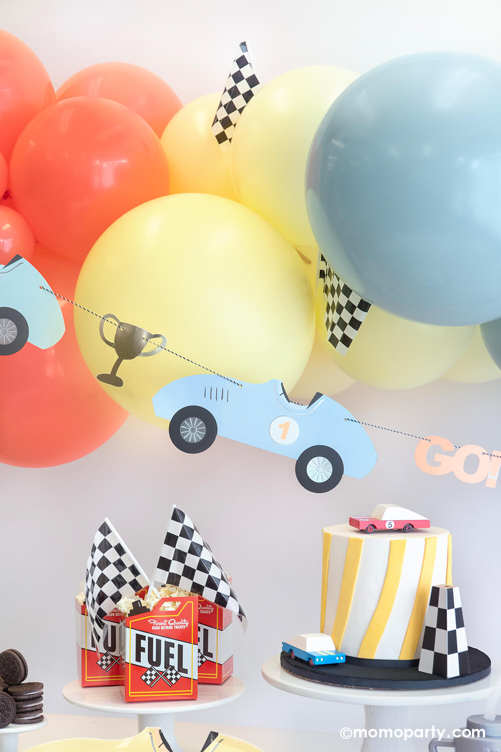 A festive race car themed party set up by Momo Party featuring a pastel balloon garland adorned with checkered flags, underneath hung a vintage race car party garland. On the table are some fuel treat boxes topped with checkered flags and a modern birthday cake topped with a race car wooden toy car toppers - a perfect inspiration for a kid's "Two Fast" second birthday party or "Fast One" first birthday party.