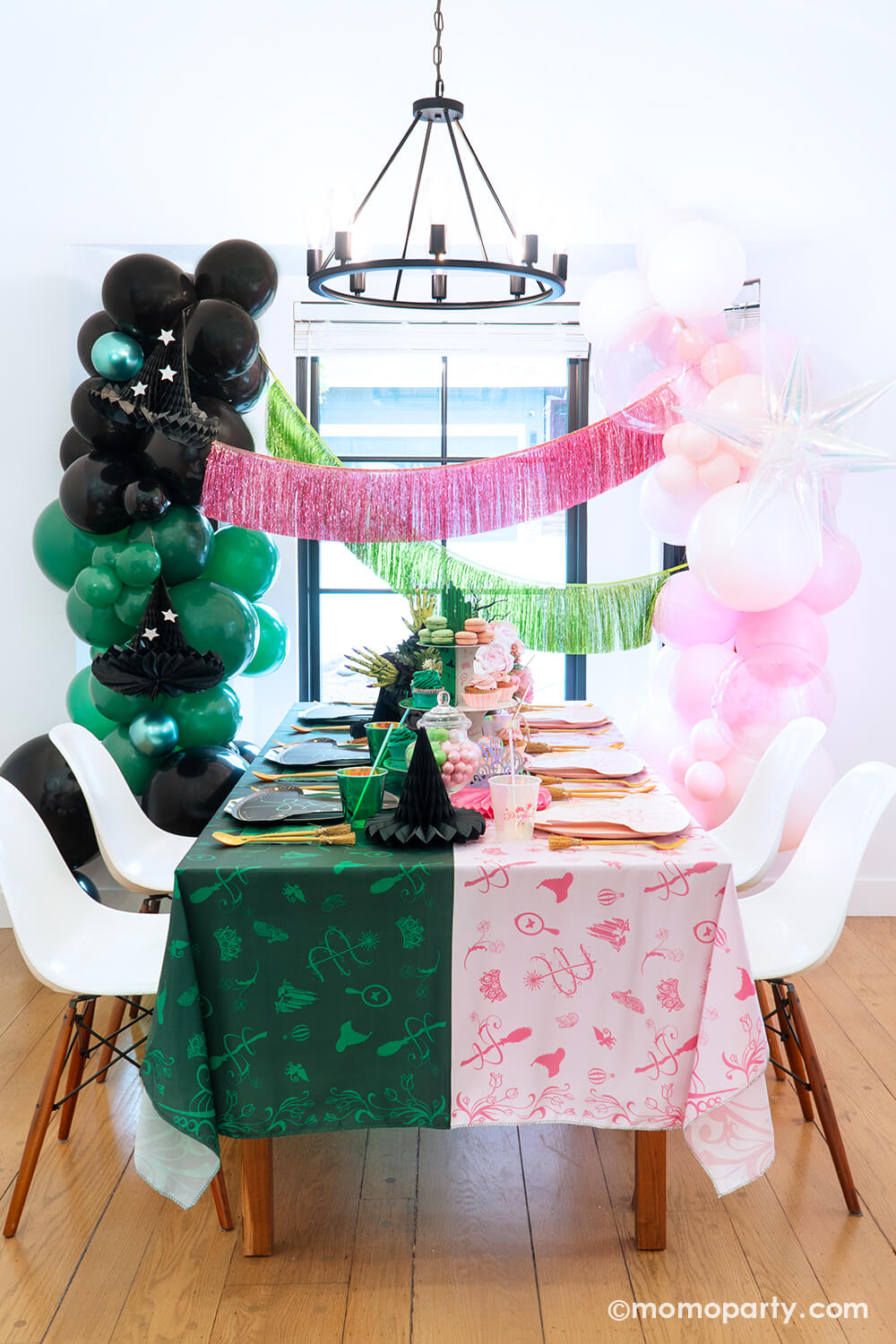 A spellbinding Wicked party set up by Momo Party featuring various Wicked themed party supplies and decorations that represent two worlds or Glinda and Elphaba including pink and black witch hat shaped plates, two-toned Emerald City cupcake stand, Wicked party cups, napkins, witch broom utensil set and Wicked witch hat honeycombs on the green and pink two-tone tablecloth. With Wicked floral arrangement and treats on the table, balloon garlands and pink and green fringe garlands hung above as the backdrop.