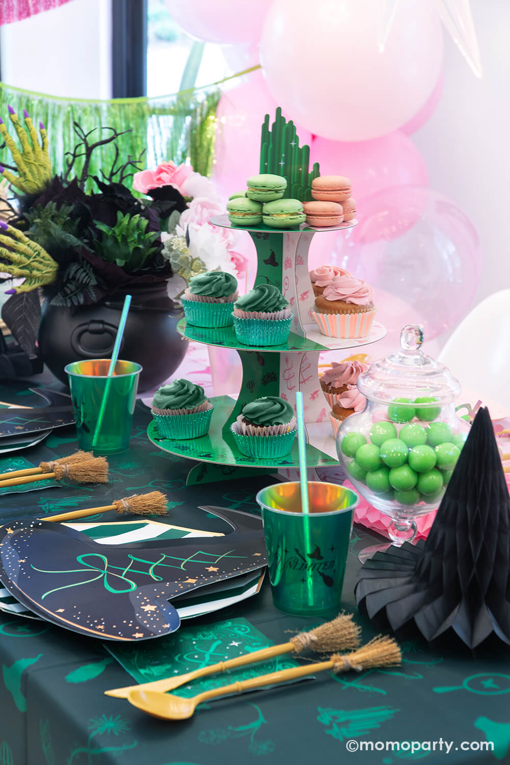A Wicked themed party table setting featuring various Wicked themed party supplies and decorations from Momo Party including black and green Wicked witch hat shaped plates, two-toned Wicked Emerald City cupcake stand, Wicked party cups, witch broom utensil set and Wicked witch hat honeycombs on the Wicked green and pink two-tone tablecloth. With black and green floral bouquet and treats, this makes a mystical yet whimsical table for a Wicked themed party in this spooky season.