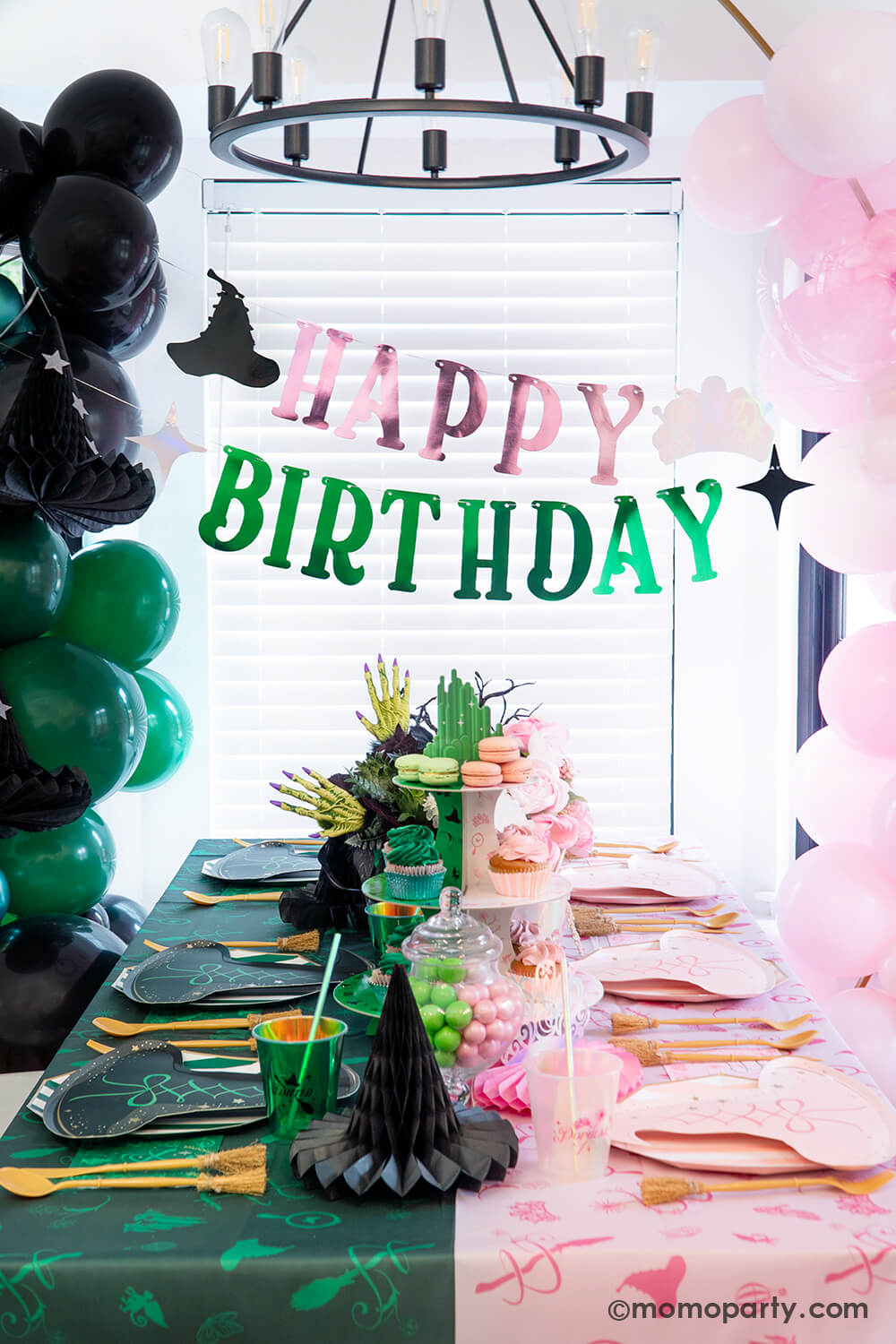A spellbinding Wicked birthday party set up by Momo Party featuring various Wicked themed party supplies and decorations that represent two worlds or Glinda and Elphaba including pink and black witch hat shaped plates, two-toned Emerald City cupcake stand, Wicked party cups, napkins, witch broom utensil set and Wicked witch hat honeycombs on the green and pink two-tone tablecloth. With Wicked floral arrangement and treats on the table, balloon garlands and happy birthday banner hung above as the backdrop.