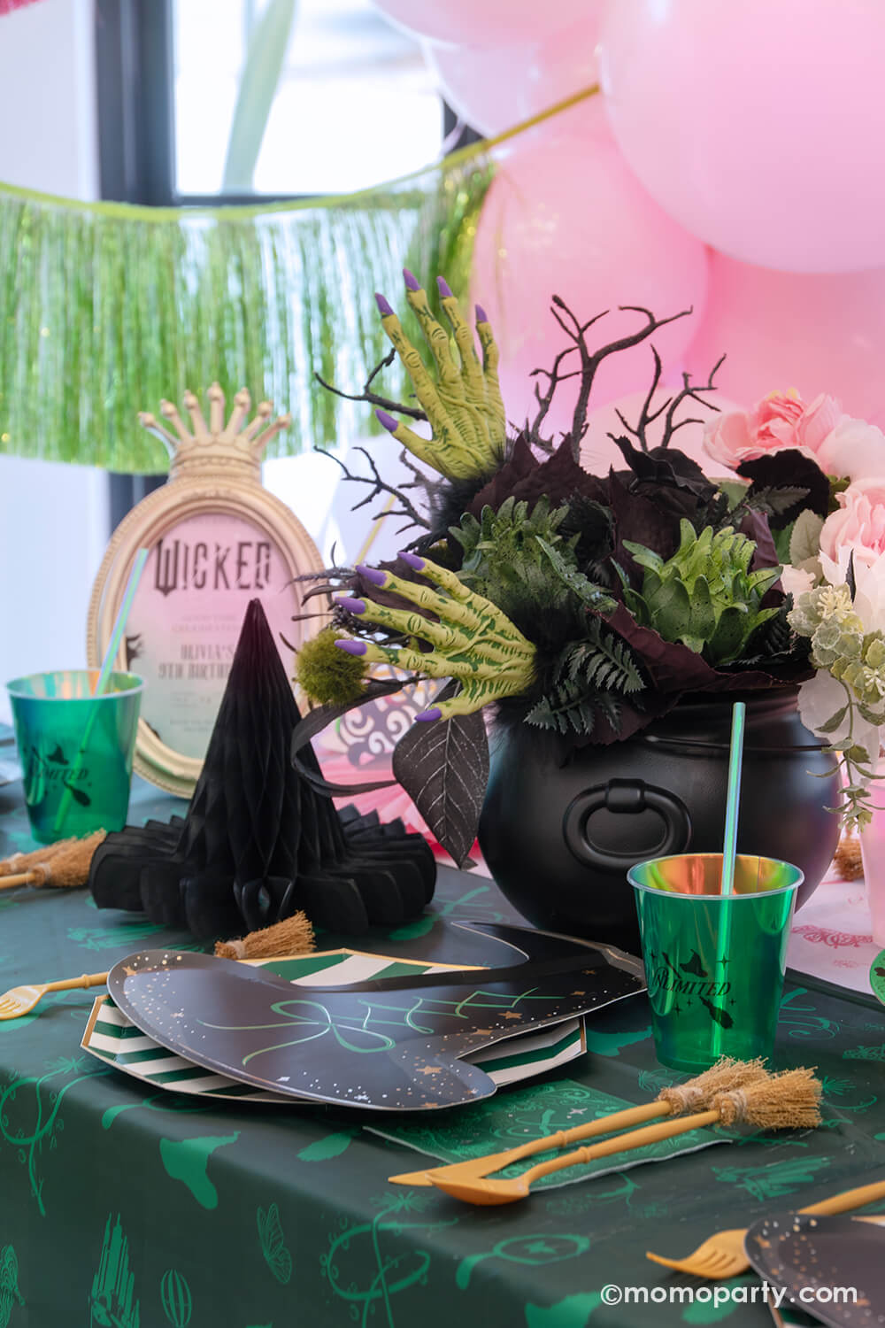 A Wicked themed party table setting with various of Momo Party's Wicked themed party supplies including the wicked witch hat shaped plates, emerald green striped dinner plates, the Wicked green napkins inspired by Elphaba, witch broom shaped utensil set, and wicked green party cups with green iridescent straws on the green and pink two-tone Wicked tablecloth. With Wicked inspired floral bouquet and witch hat honeycombs, it's perfect for a spellbinding celebration.