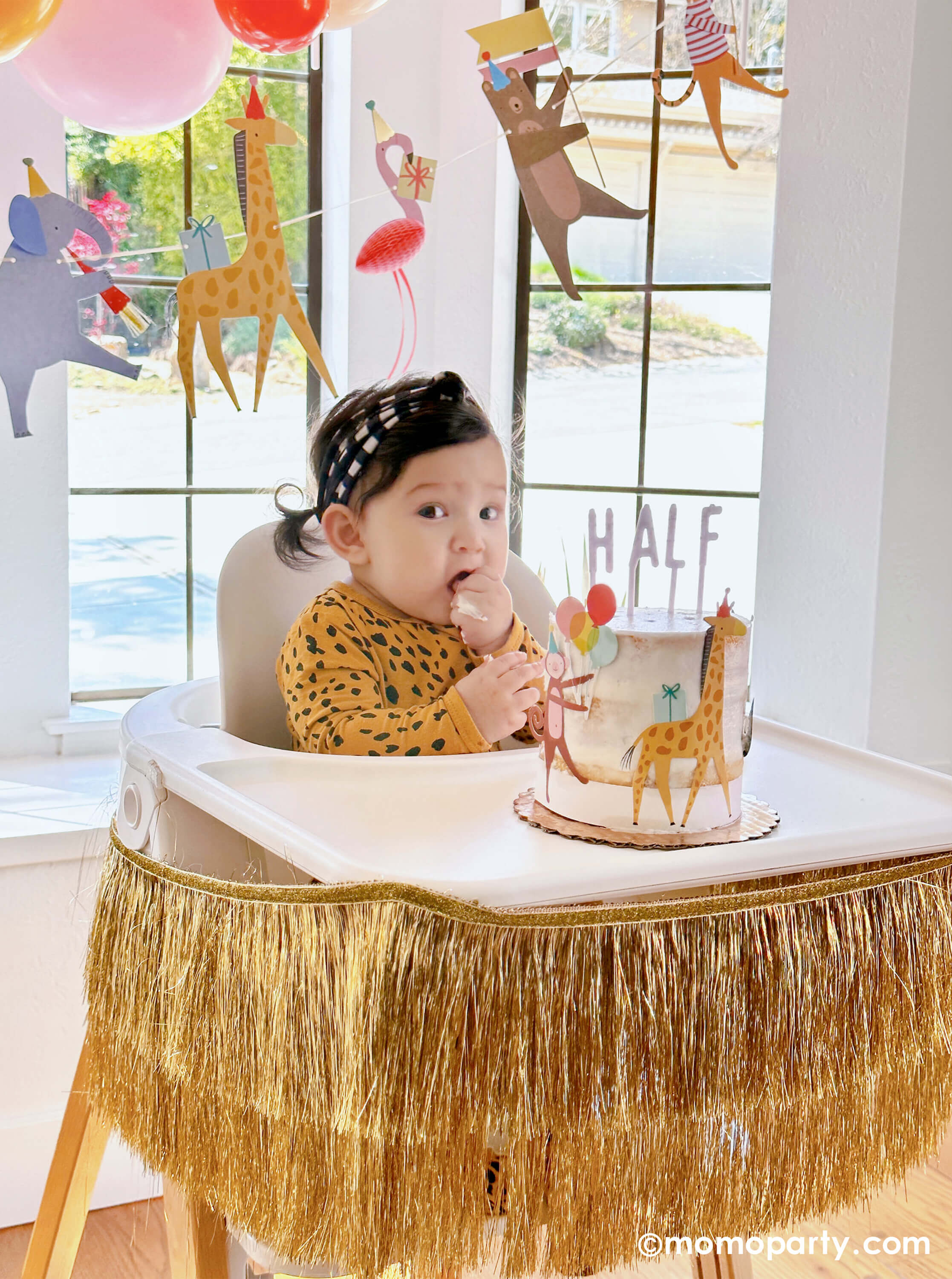 A cute baby girl wearing animal pattern onesie, sitting on a highchair in her 6 months animal themed half birthday party, eating her Naked Cake decorated with Meri Meri Animal Parade Cake Wrap & Toppers, and on the top spelling "HALF" with Cake By Courtney Letter-board Cake Toppers, repersend for a six months half birthday, along with Meri Meri gold fringe garland around the highchair tray, it makes a great party inspiration for kid's animal, safari, carnival themed birthday party.