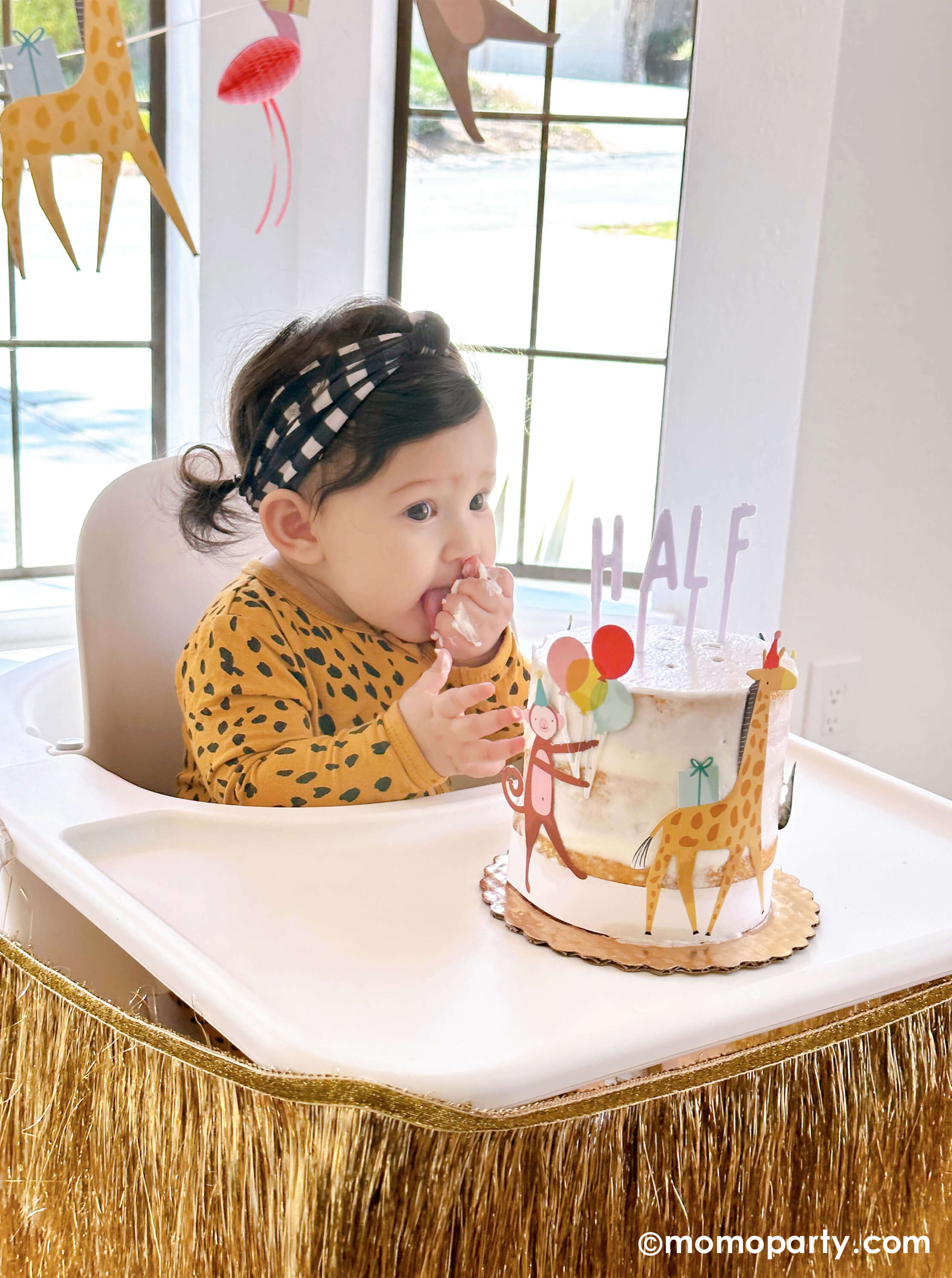  A cute baby girl wearing animal pattern onesie, sitting on a highchair in her 6 months animal themed half birthday party, eating her Naked Cake decorated with Meri Meri Animal Parade Cake Wrap & Toppers, and on the top spelling "HALF" with Cake By Courtney Letter-board Cake Toppers, repersend for a six months half birthday, along with Meri Meri gold fringe garland around the highchair tray, it makes a great party inspiration for kid's animal, safari, carnival themed birthday party.