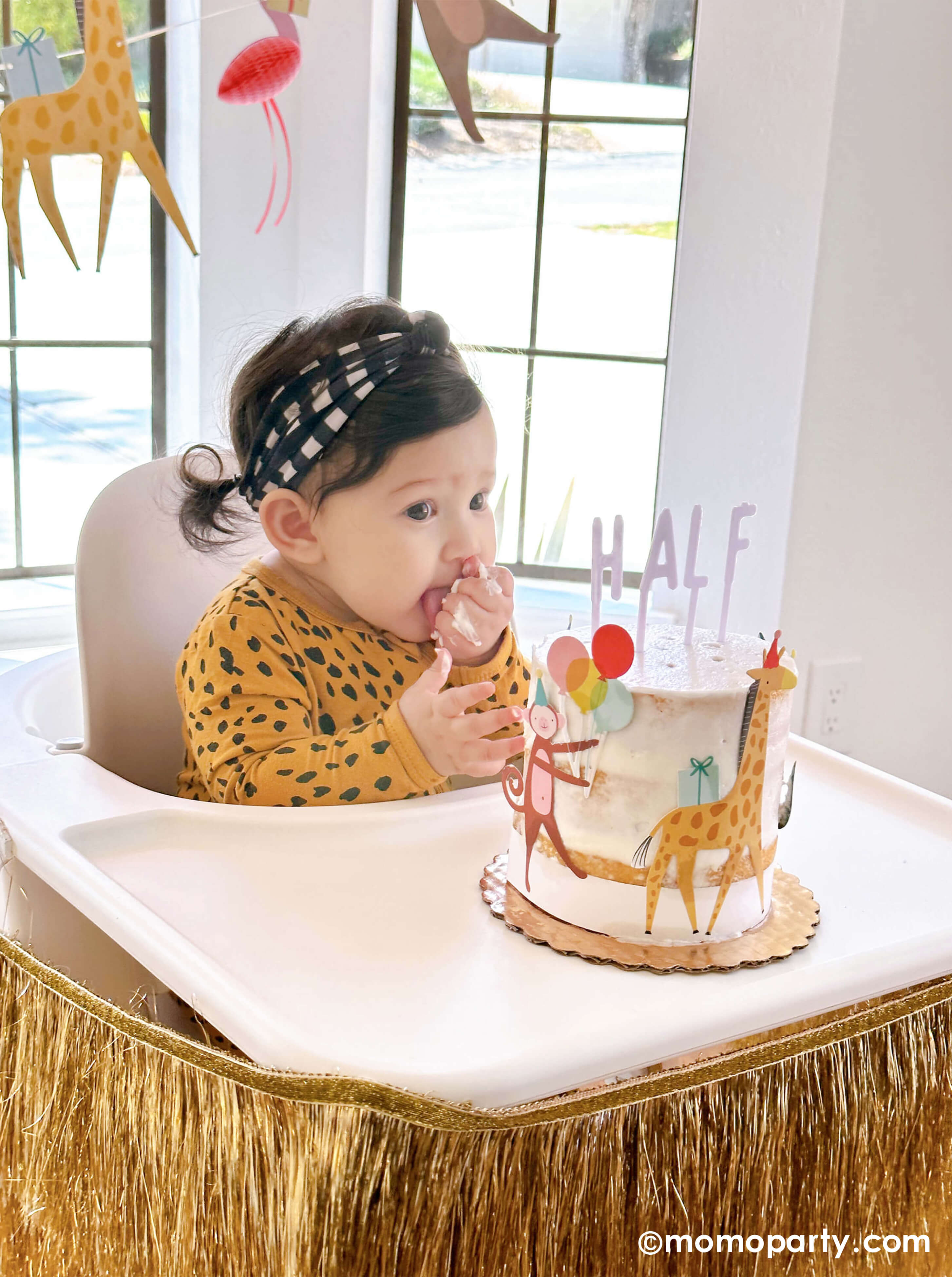 A cute baby girl wearing animal pattern onesie, sitting on a highchair in her 6 months animal themed half birthday party, eating her Naked Cake decorated with Meri Meri Animal Parade Cake Wrap & Toppers, and on the top spelling "HALF" with Cake By Courtney Letter-board Cake Toppers, repersend for a six months half birthday, along with Meri Meri gold fringe garland around the highchair tray, it makes a great party inspiration for kid's animal, safari, carnival themed birthday party.