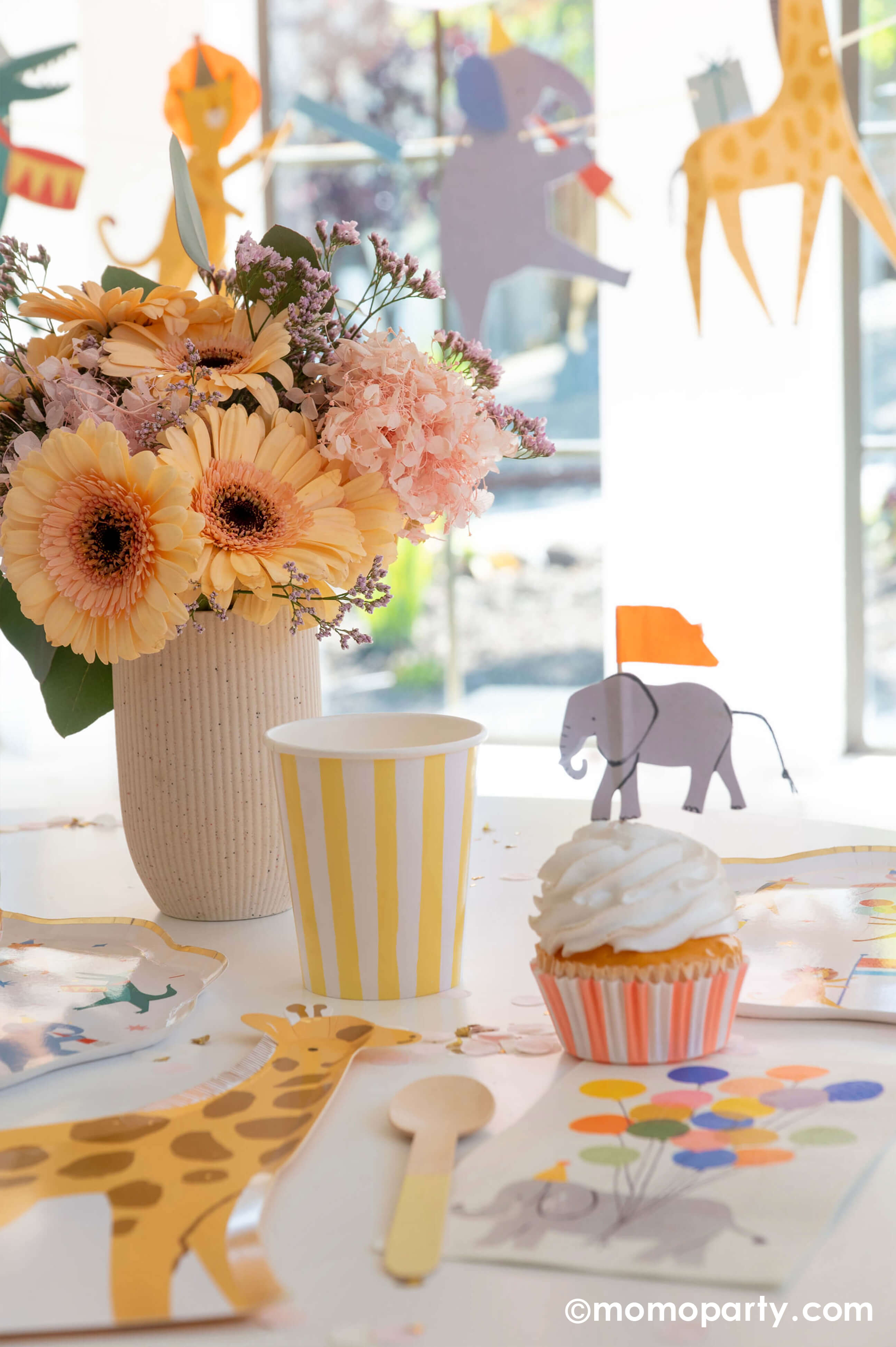 A kid's animal themed party table featuring Momo Party's Animal Parade Dinner plate, a giraffe shaped plate for cakes, and adorable safari animal cupcake toppersby Meri Meri, and Rifle Paper Co's party animal guest napkins with a cute illustration of an elephant with a party hat and bunch of festive balloons, along with light yellow striped cups and wooden utensil, it makes a great party inspiration for kid's animal, safari, carnival themed birthday party.