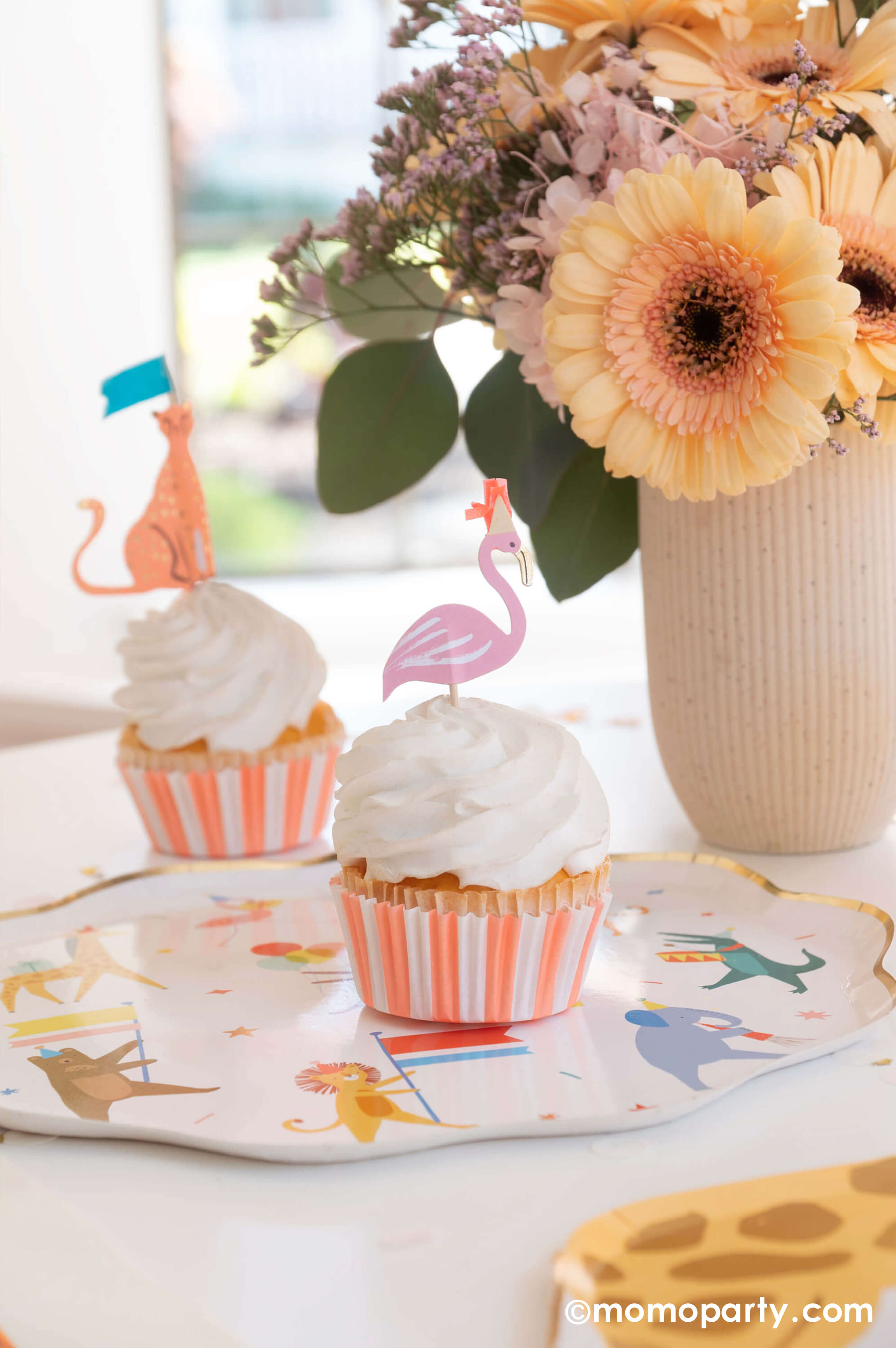 A kid's animal themed party table featuring Momo Party's Animal Parade Dinner plate, a giraffe shaped plate for cakes, and adorable safari animal cupcake toppersby Meri Meri, and Rifle Paper Co's party animal guest napkins with a cute illustration of an elephant with a party hat and bunch of festive balloons, along with light yellow striped cups and wooden utensil, it makes a great party inspiration for kid's animal, safari, carnival themed birthday party.