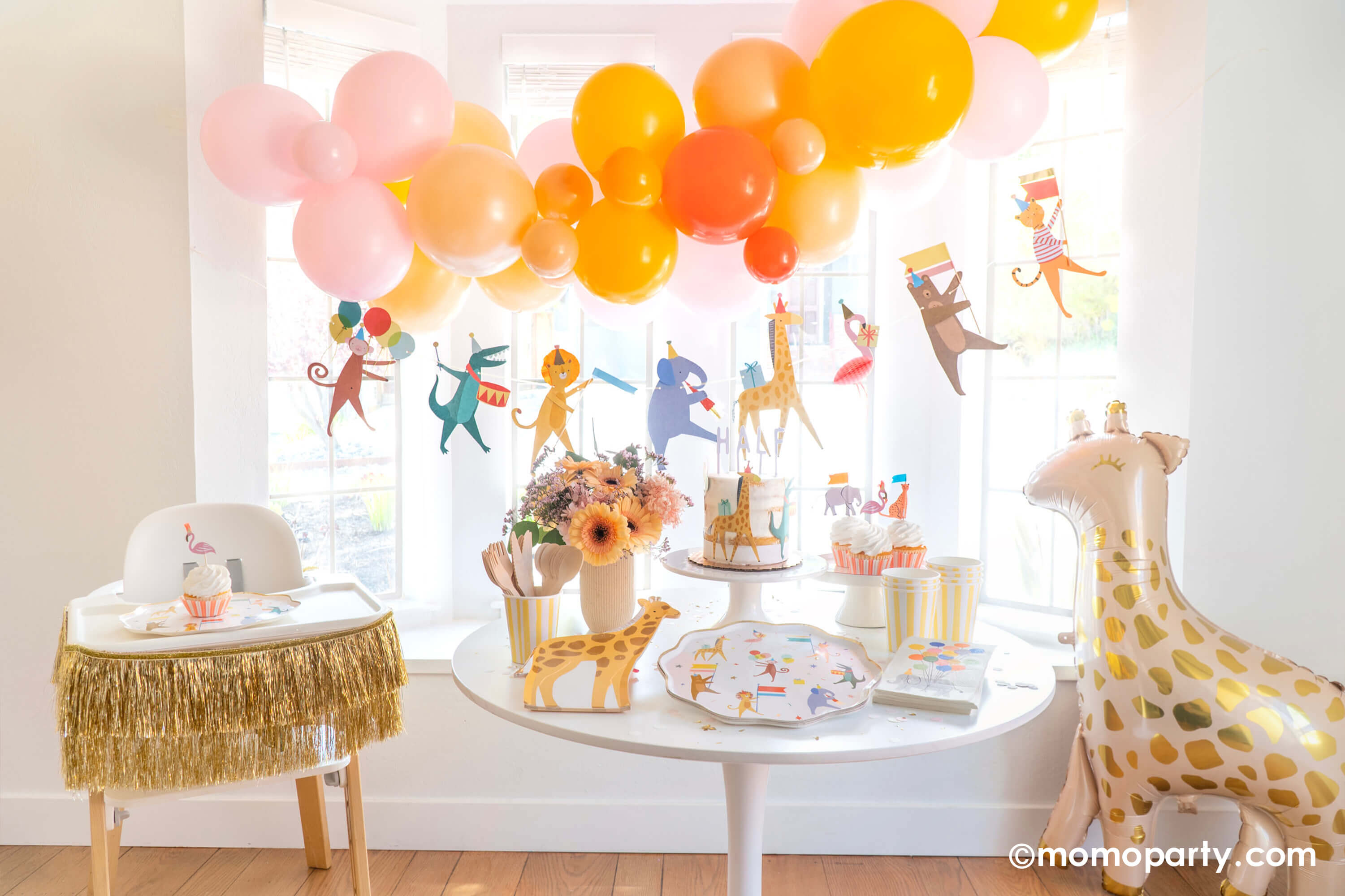 Adorable baby's animal themed party featuring Momo Party's animal parade birthday garland featuring a lion, an elephant, an alligator, a giraffe, along with a festive balloon garland in a warm color tone hanging above. On the dessert table, there are Meri Meri's animal parade dinner plates, giraffe shaped plates and cupcakes dressed with the safari animal toppers, next to the table there's a giraffe shaped foil balloon. One the left a highchair is decorated with a gold tinsel garland.