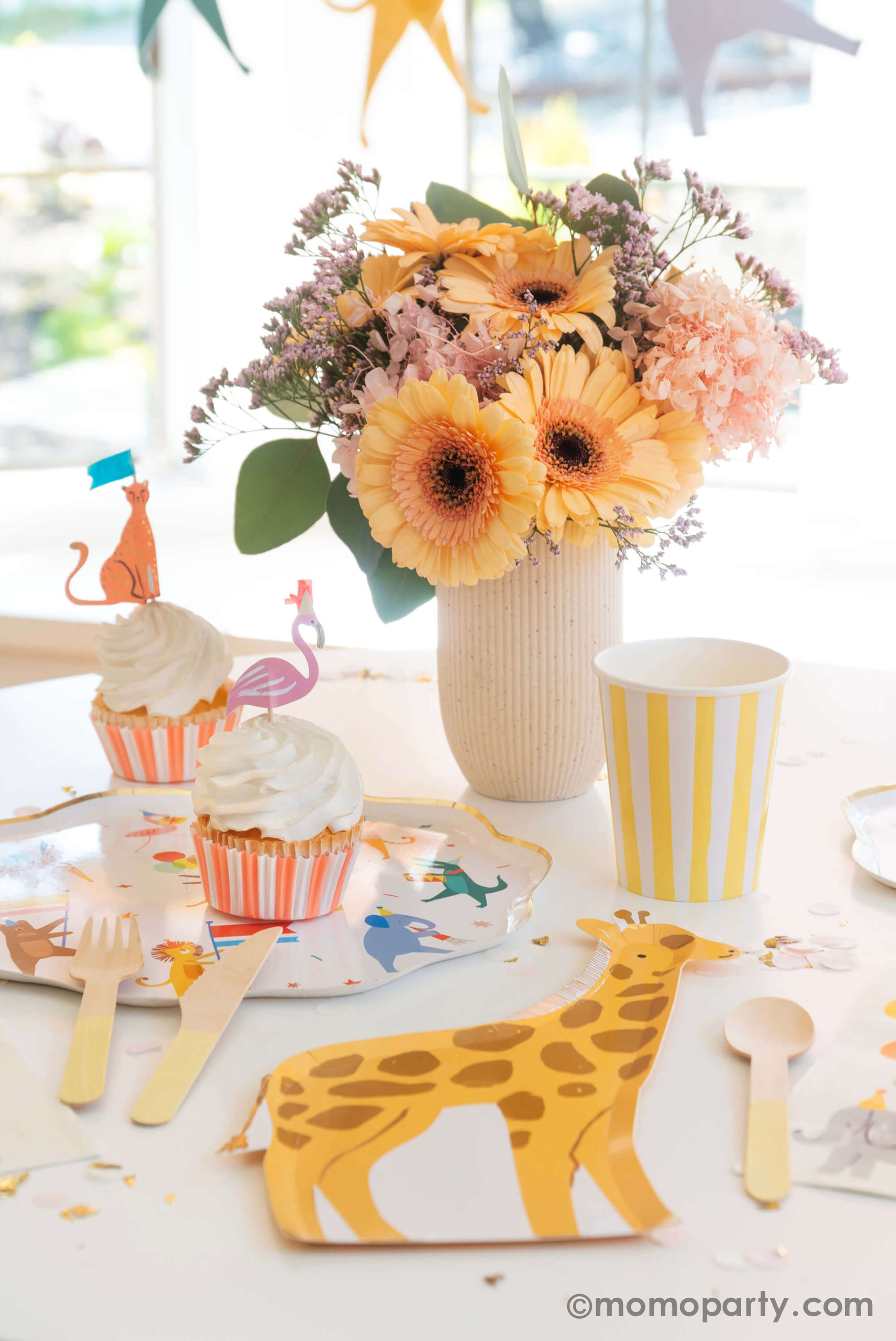 A kid's animal themed party table featuring Momo Party's Animal Parade Dinner plate, a giraffe shaped plate for cakes, and adorable safari animal cupcake toppersby Meri Meri, and Rifle Paper Co's party animal guest napkins with a cute illustration of an elephant with a party hat and bunch of festive balloons, along with light yellow striped cups and wooden utensil, it makes a great party inspiration for kid's animal, safari, carnival themed birthday party.