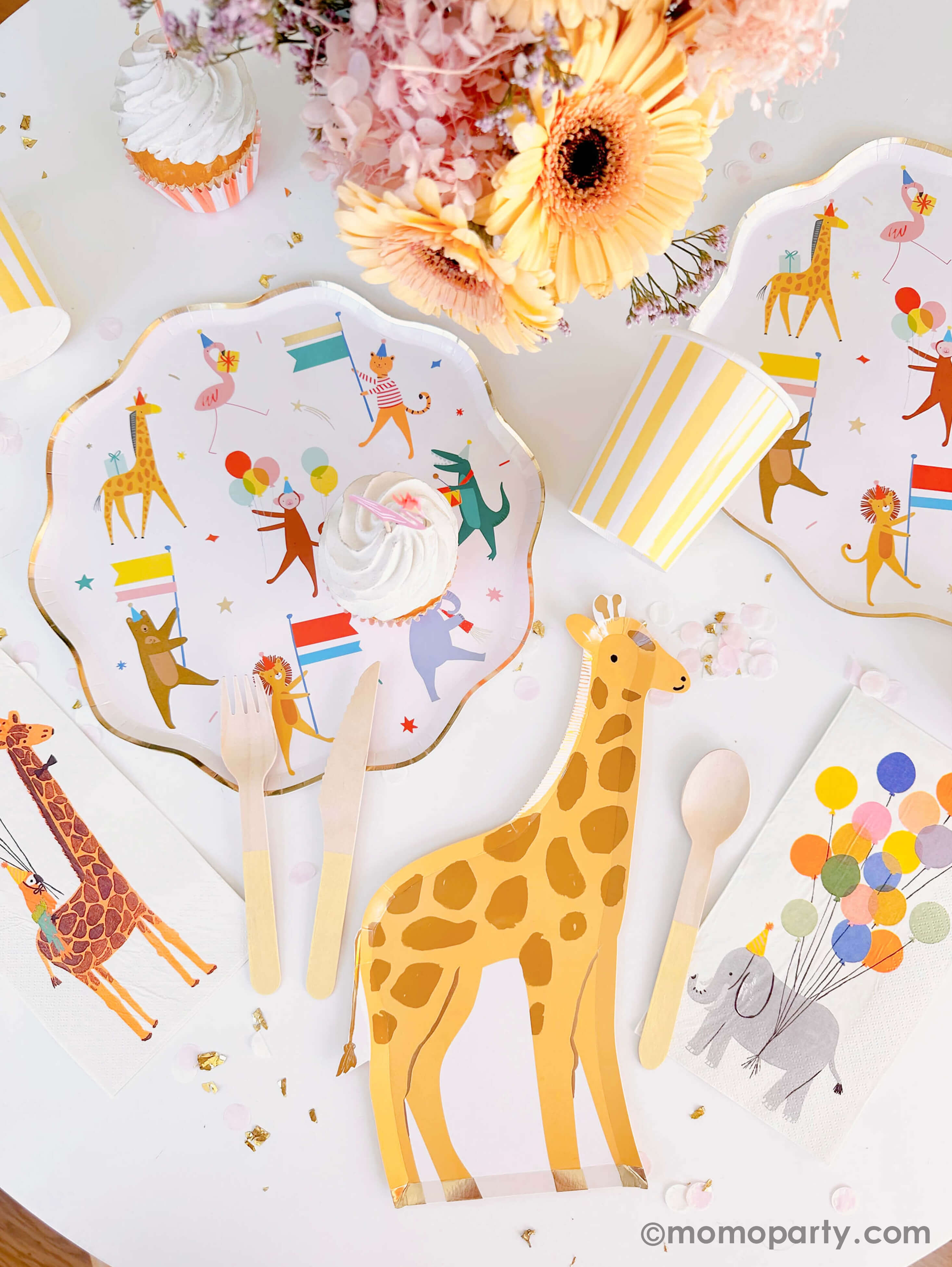 A kid's animal themed tablescape featuring Momo Party's Animal Parade Dinner plate with gold scallop edge and cute animal illustration designs, a giraffe shaped plate for cakes by Meri Meri, and Rifle Paper Co's party animal guest napkins with a cute illustration of an elephant with a party hat and bunch of festive balloons, along with light yellow striped cups and wooden utensil, it makes a great party inspiration for kid's animal, safari, carnival themed birthday party.