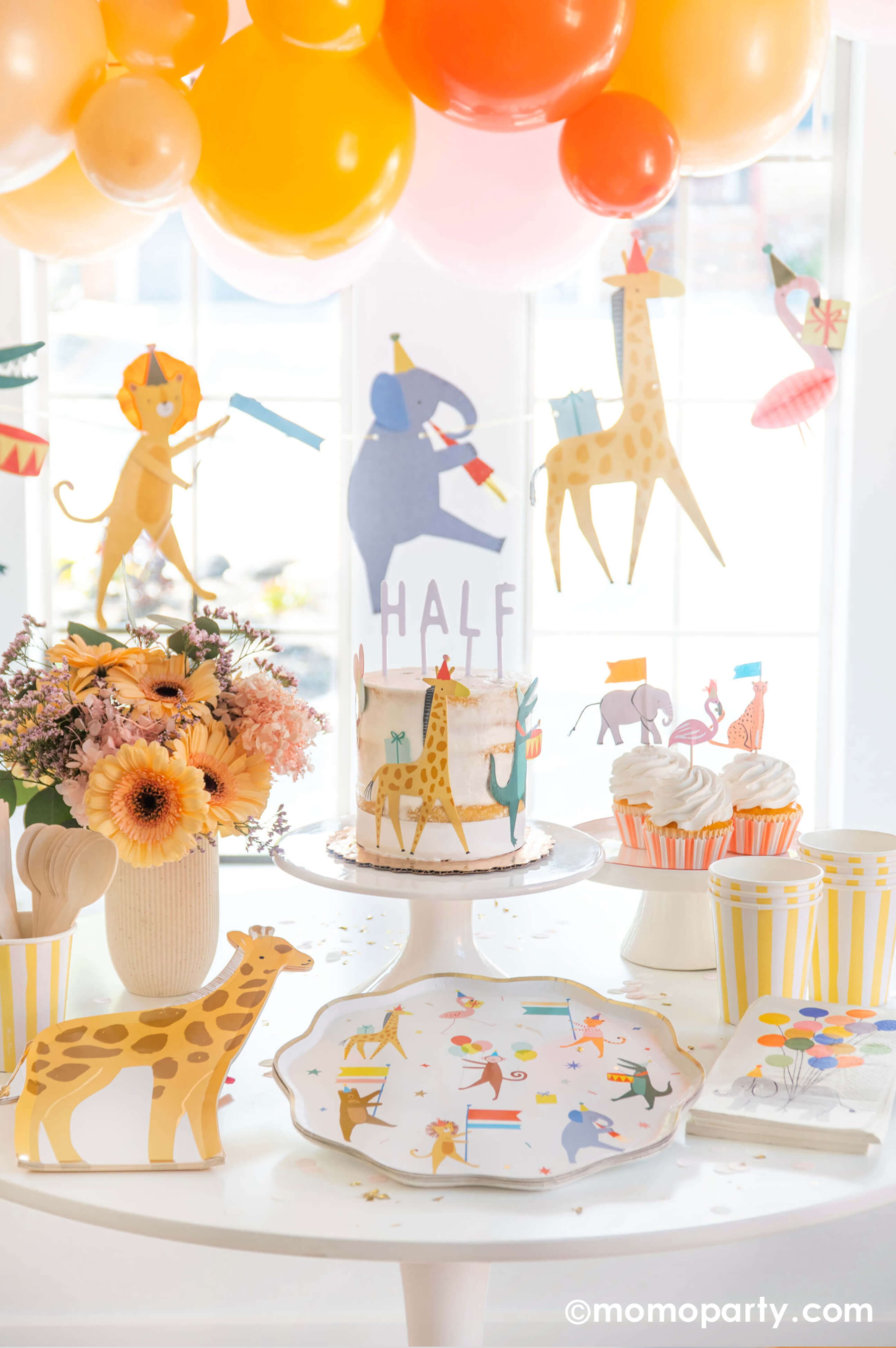 A kid's animal themed party table featuring Momo Party's Animal Parade Dinner plate, giraffe shaped plates, light yellow striped cups and safari animal cake toppers by Meri Meri, along with a beautiful spring flower arrangement as the centerpiece and a fun animal parade birthday garland featuring a lion, an elephant, a giraffe, a flamingo, and a festive balloon garland in a warm color tone in the back, it makes a great party inspiration for kid's animal, safari, carnival themed birthday party.