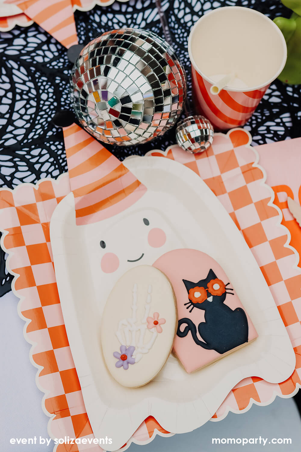 Details of Momo Party Groovy Halloween Themed Party Table, featuring a cookie with black cat and skeleton hand design on the My Mind's Eye Ghoul Gang Ghost Shaped Paper Plates, laid over Ghoul Gang Checkered Paper Plates, Iridescent Party Straws inside the Meri Meri Pink & Orange Stripy Cups, Shining Disco balls, They all on the black spider web placemats. This is such a modern fun Halloween theme and Halloween party for kids.