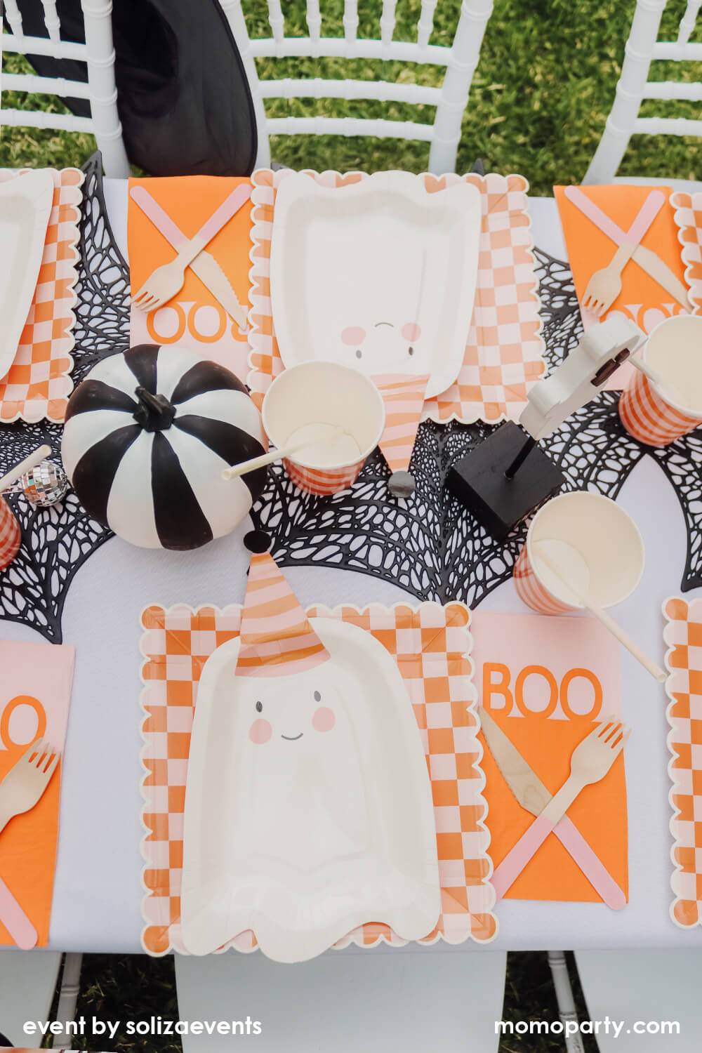 Top view of Momo Party Groovy Halloween Themed Party Table, decorated with 6 sets of My Mind's Eye Ghoul Gang Ghost Shaped Paper Plates, over Ghoul Gang Checkered Paper Plates, Happy Haunting Boo Guest Towels, Iridescent Party Straws inside the Meri Meri Pink & Orange Stripy Cups, They all on the black spider web placemats, and there is a black and white striped pumpkin, a cute ghost Ghost Cutout with a Boo base as the centerpiece. This is such a modern fun Halloween theme and Halloween party for kids.