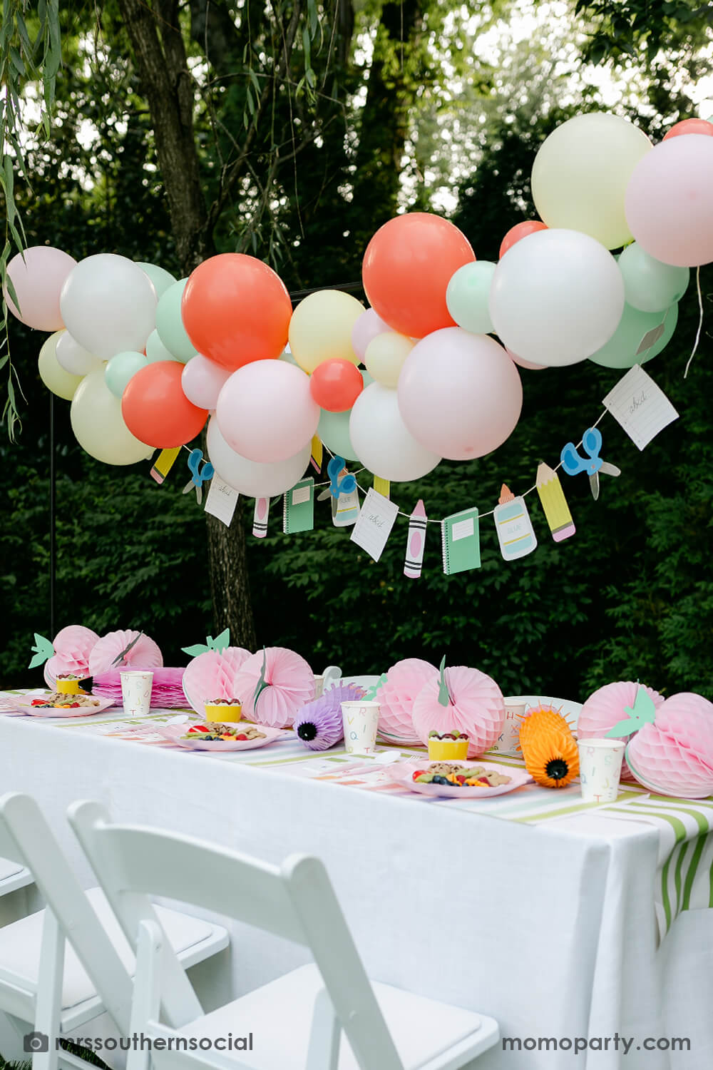 A backyard pastel themed back to school party table set up and decoration featuring Momo Party's back to school themed party goods including back to school party garland, pink apple honeycombs and rainbow pastel pencil honeycombs as the centerpiece, ABC alphabet plates, party cups and napkins. With a pastel themed balloon garland hung above the table, it makes an adorable celebration for kid's first day of school.