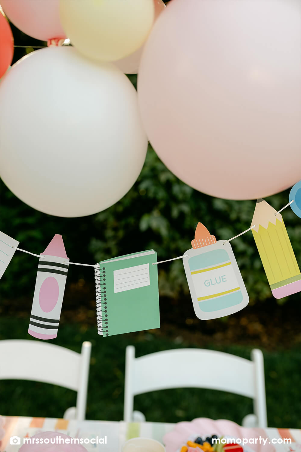 A backyard pastel themed back to school party decorated with Momo Party's back to school themed party goods including back to school party garland featuring school supply pennants including pencils, glue bottles, crayons, notebooks,  and more. With a pastel themed balloon garland hung above the table, it makes an adorable celebration for kid's first day of school.
