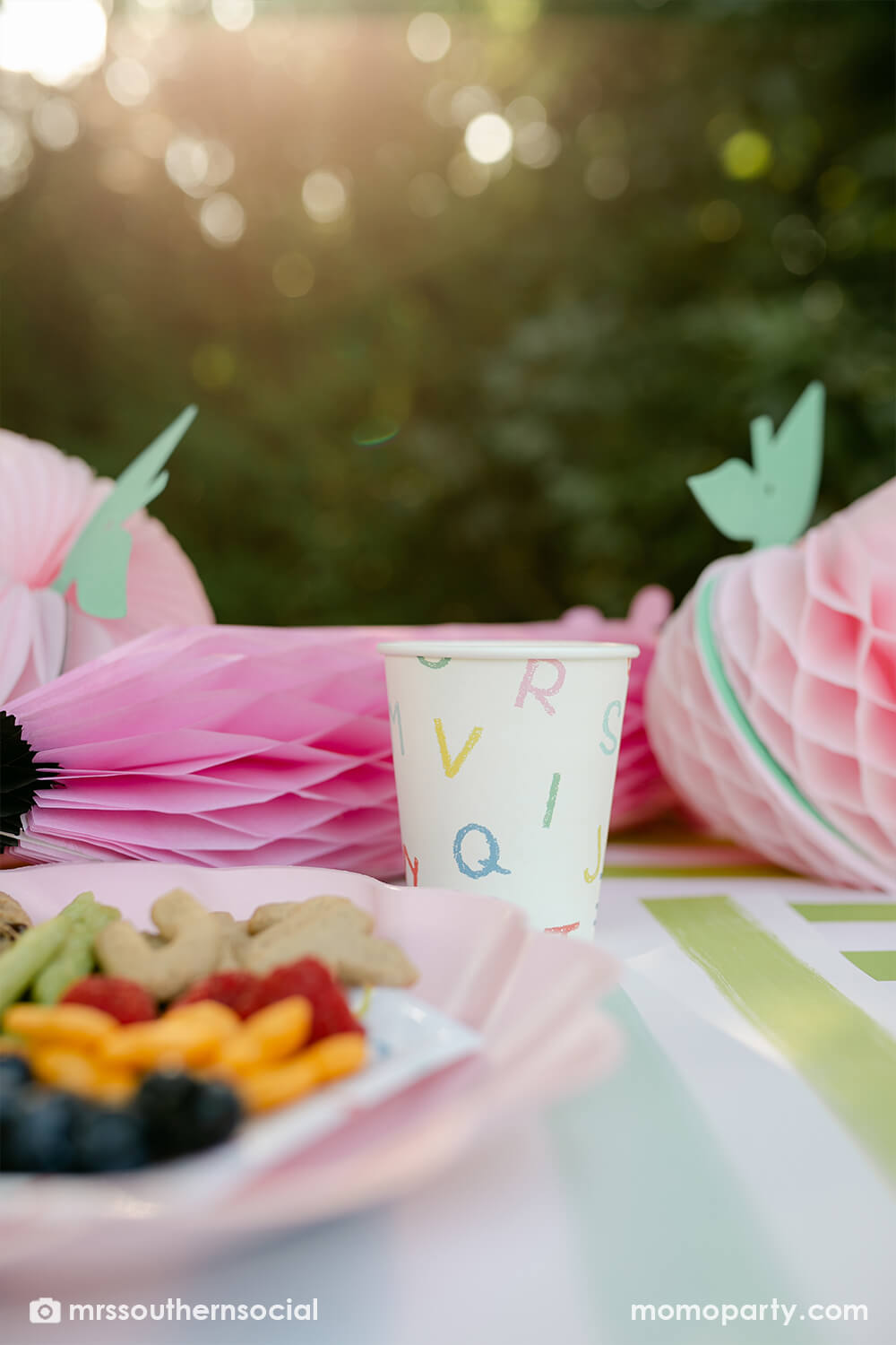 A backyard pastel back to school party table featuring Momo Party's back to school party supplies including ABC alphabet plates, napkins, party cups by Merrilulu. With pastel rainbow striped placemats and pastel rainbow pencil honeycombs and pink apple honeycombs, all together makes an adorable tablescape for kid's first day of school celebration.