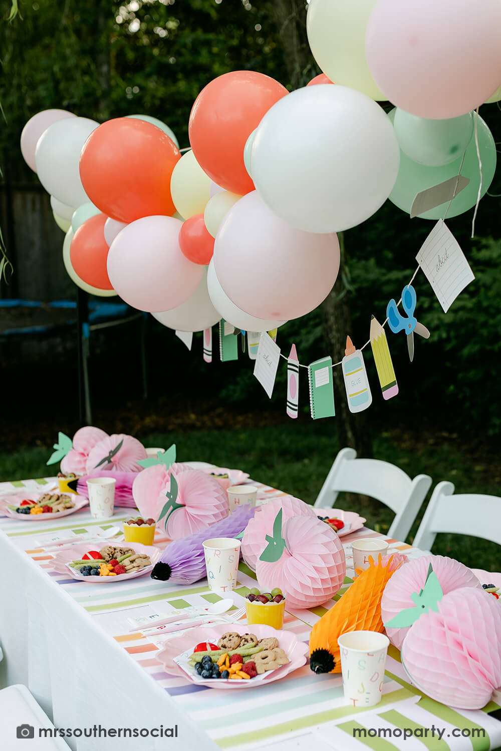A backyard pastel themed back to school party table set up and decoration featuring Momo Party's back to school themed party goods including back to school party garland, pink apple honeycombs and rainbow pastel pencil honeycombs as the centerpiece, ABC alphabet plates, party cups and napkins. With a pastel themed balloon garland hung above the table, it makes an adorable celebration for kid's first day of school.