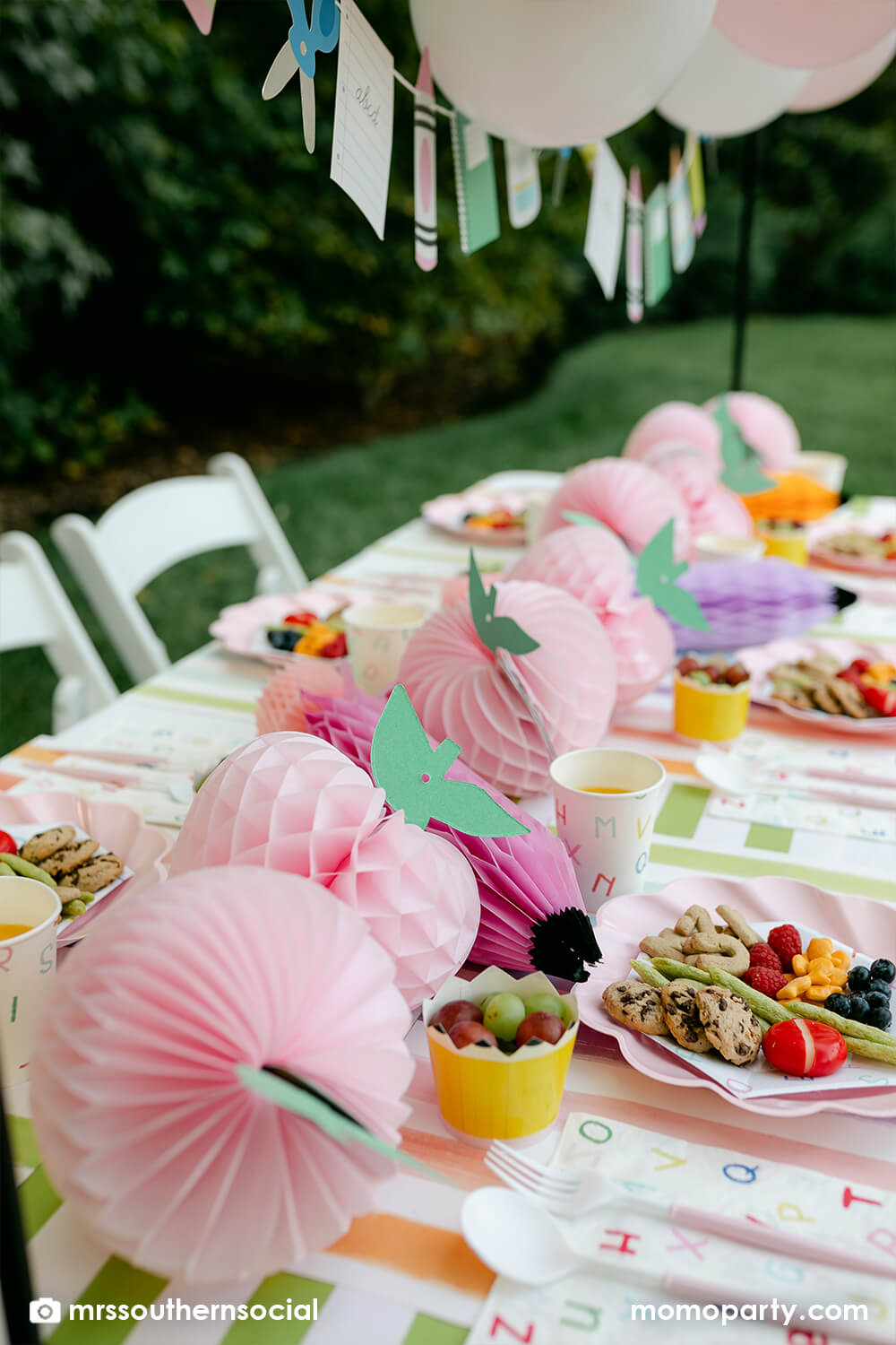A backyard pastel themed back to school party table set up and decoration featuring Momo Party's back to school themed party goods including back to school party garland, pink apple honeycombs and rainbow pastel pencil honeycombs as the centerpiece, ABC alphabet plates, party cups and napkins. With kids snacks including ABC alphabet crackers, fruits, cheese, it gives great ideas and inspo for celebrating kid's first day of school.