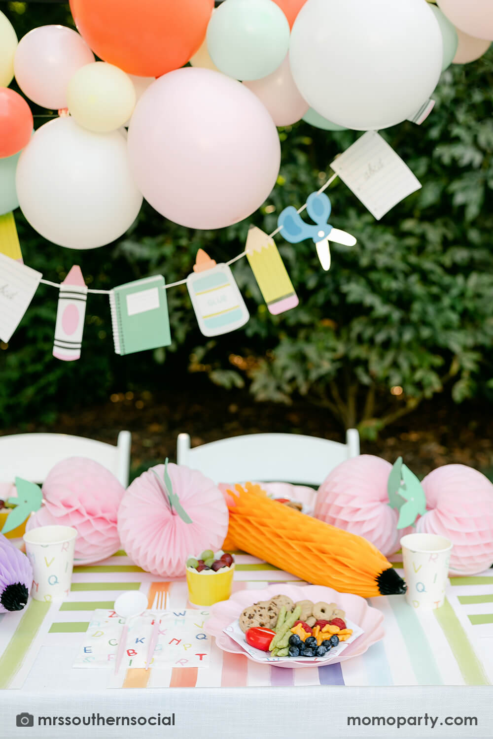 A backyard pastel themed back to school party table set up and decoration featuring Momo Party's back to school themed party goods including back to school party garland, pink apple honeycombs and rainbow pastel pencil honeycombs as the centerpiece, ABC alphabet plates, party cups and napkins. With a pastel themed balloon garland hung above the table, it makes an adorable celebration for kid's first day of school.
