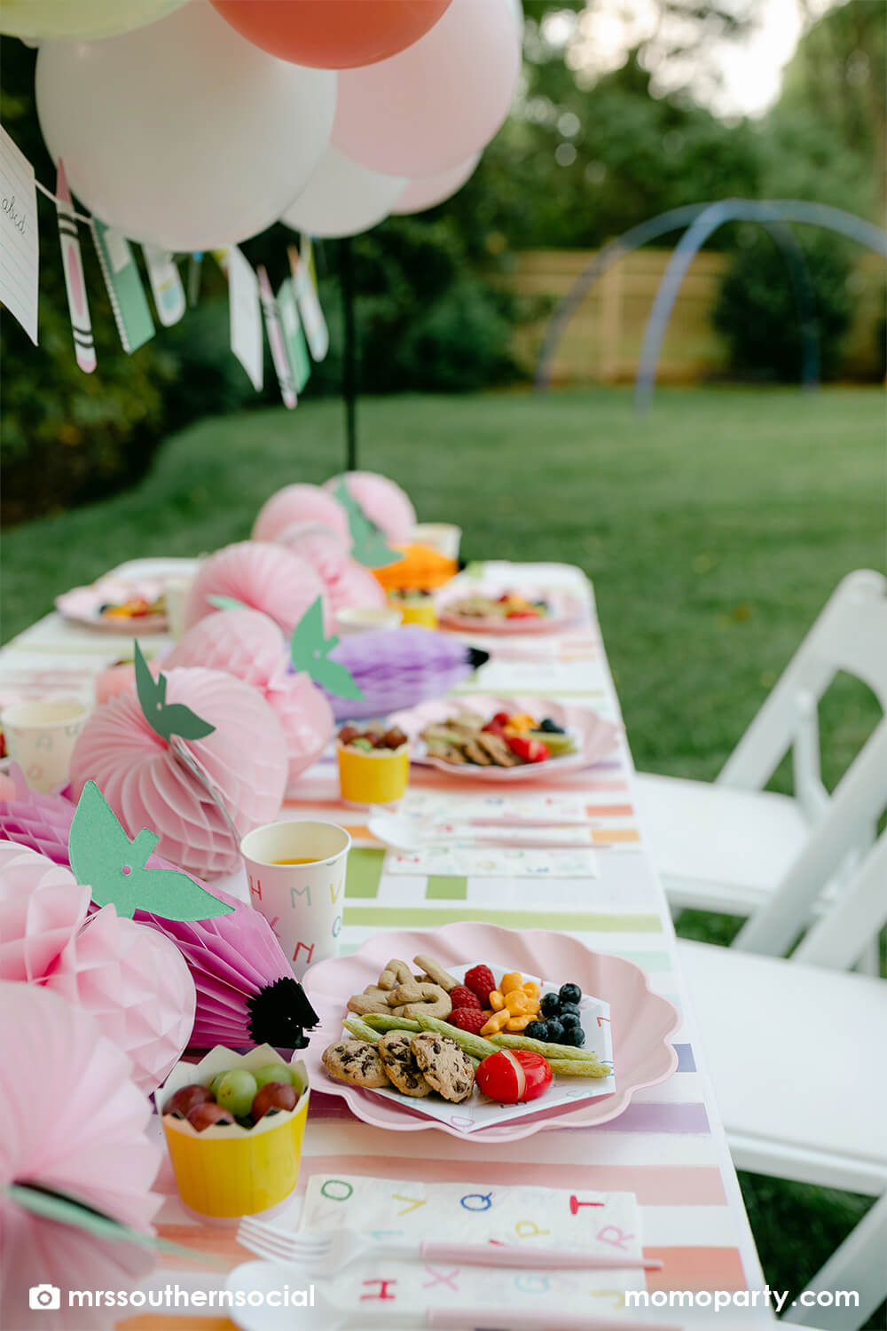 A pastel back to school party table featuring Momo Party's back to school party supplies including ABC alphabet plates, napkins party cups, and pencil shaped food cups. One the plates are kids snack ideas including fruits, cheeses, goldfish, ABC alphabet crackers. With pastel rainbow striped placemats and pastel rainbow pencil honeycombs and pink apple honeycombs, all together makes an adorable tablescape for kid's first day of school celebration.