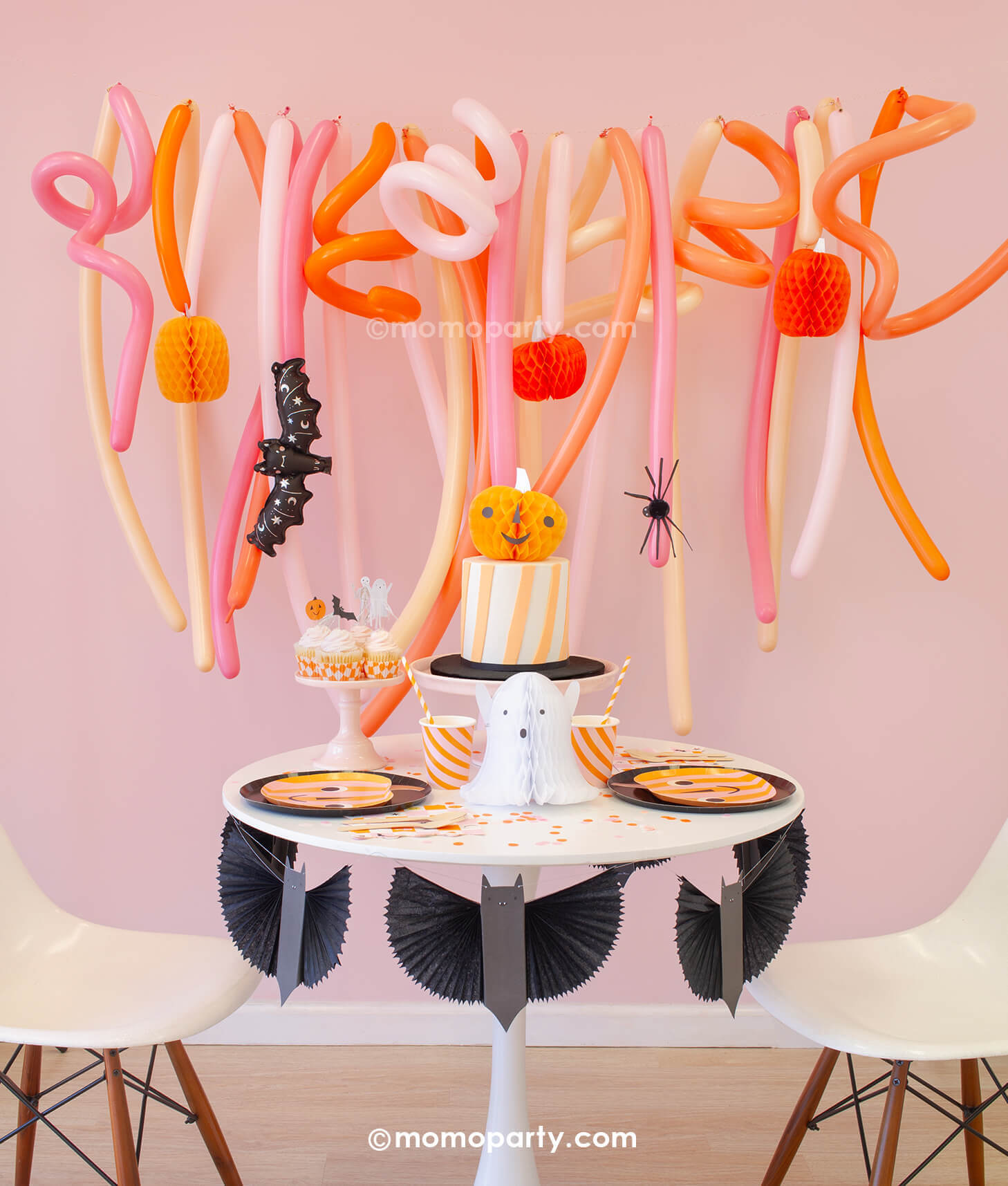 Momo Party's Trick or Treat Pink Halloween box set up featuring a festive long balloon backdrop with curling long balloons  in coral, pink, rose and peach colors. On the balloon backdrop it's decorated with paper pumpkin honeycombs and a mini bat foil balloon. In front of the balloons, a small round table is filled with Halloween party supplies including pumpkin shaped plates, a bat garland, ghost and pumpkin honeycomb decorations from momoparty.com.