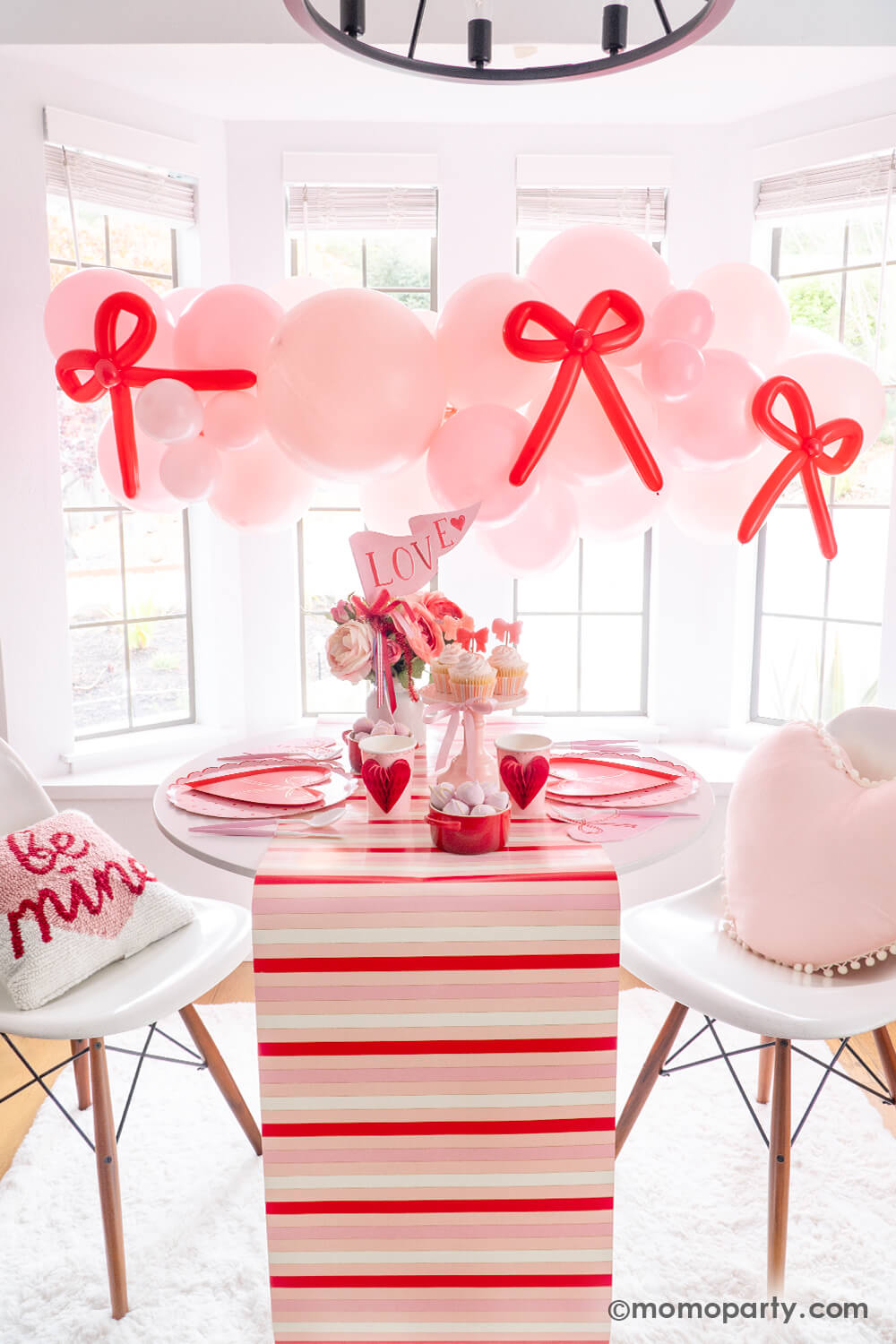 Tying the Knot of Love: A Bow-Themed Valentine's Day Party Set up by Momo Party. A pink balloon garland with a red bow-shaped balloon hangs above the Bow-tastic Table, adorned with Meri Meri Heart Pattern Dinner Plates, Bow Plates, matching napkins, Honeycomb Heart Cups, Blush and White Cutlery Set, Pink Bow Candles on cupcakes, and a Love Party Pennant gracing the flower vase. All elegantly arranged on a red-striped paper runner. A cute idea for your Valentine's celebration or Galentine's Day with besties