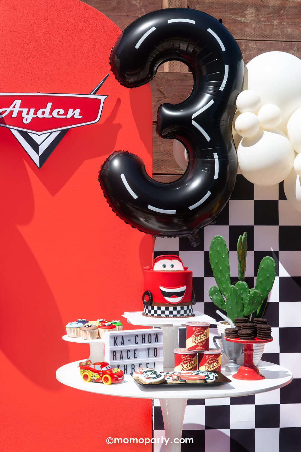 A kid's Disney Cars Lightning Mcqueen themed birthday party cake table decoration featuring various race car themed party supplies form Momo Party's including the Winner Circle race car felt party pennant, the trophy shaped tumbler, the tire shaped sipper, and fuel gas tin shaped treat boxes. In the background, there's the number 3 road tape foil balloon floating above, along with the custom-made Disney Cars logo and the checkered background, this festive decoration is perfect for boys birthday party!