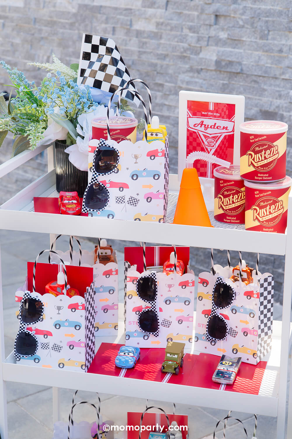 A kid's Disney Cars themed birthday party favor cart with various race car themed party favors and bags from Momo Party including customized Disney Cars toy cars, checkered race car favor bags by Meri Meri, Rusteze potato chips cans, construction cone shaped cups, checkered flags and checkered sunglasses, making his a perfect idea for for boy's race car themed birthdays or Lightning Mcqueen themed parties.