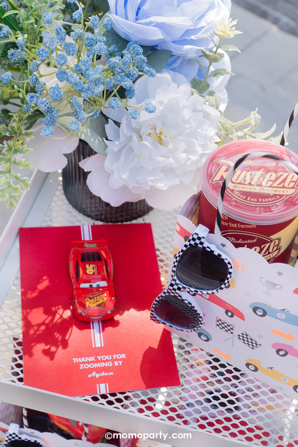 A Disney Cars Lightning Mcqueen themed birthday party red car party favor card with a Lightning Mcqueen toy car and checkered sunglasses, next to them is the Rusteze gas can of potato chips in the race car themed party favor bag by Meri Meri on a decorative table with flowers.