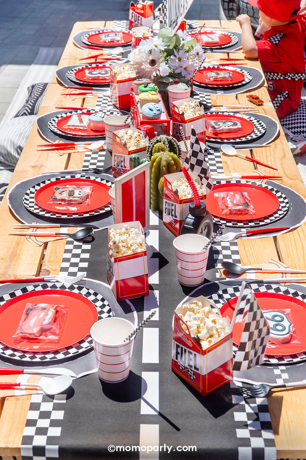 A kid's Disney Cars Lightning Mcqueen themed birthday party table setting  by Momo Party featuring a table filled with race car themed tableware including race track checkered table runner, fuel treat boxes filled with popcorn, red striped cups, red and white utensils, red and checkered plates and mini race car flags as decoration. With cactus plants, flowers and a red truck filled with cupcakes in Disney Cars designs, this kid's table setting is a perfect inspo for kid's race car themed birthday party.