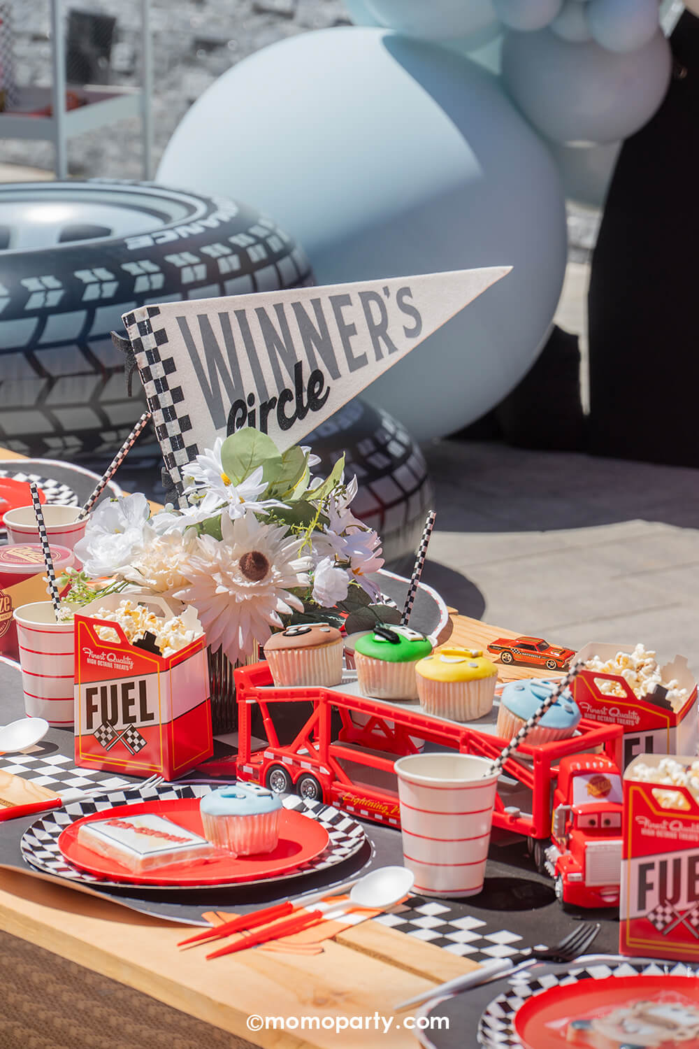 A kid's Disney Cars Lightning Mcqueen themed birthday party table set up by Momo Party featuring various race car themed party supplies including the Winner Circle race car felt party pennant, fuel treat boxes, checkered dinner plates, red striped party cups and cone shaped napkins. There's the truck 'Mack' holding Cars themed cupcakes as the centerpiece. Along with balloon decorations and tire inflatables in the back as the backdrop.
