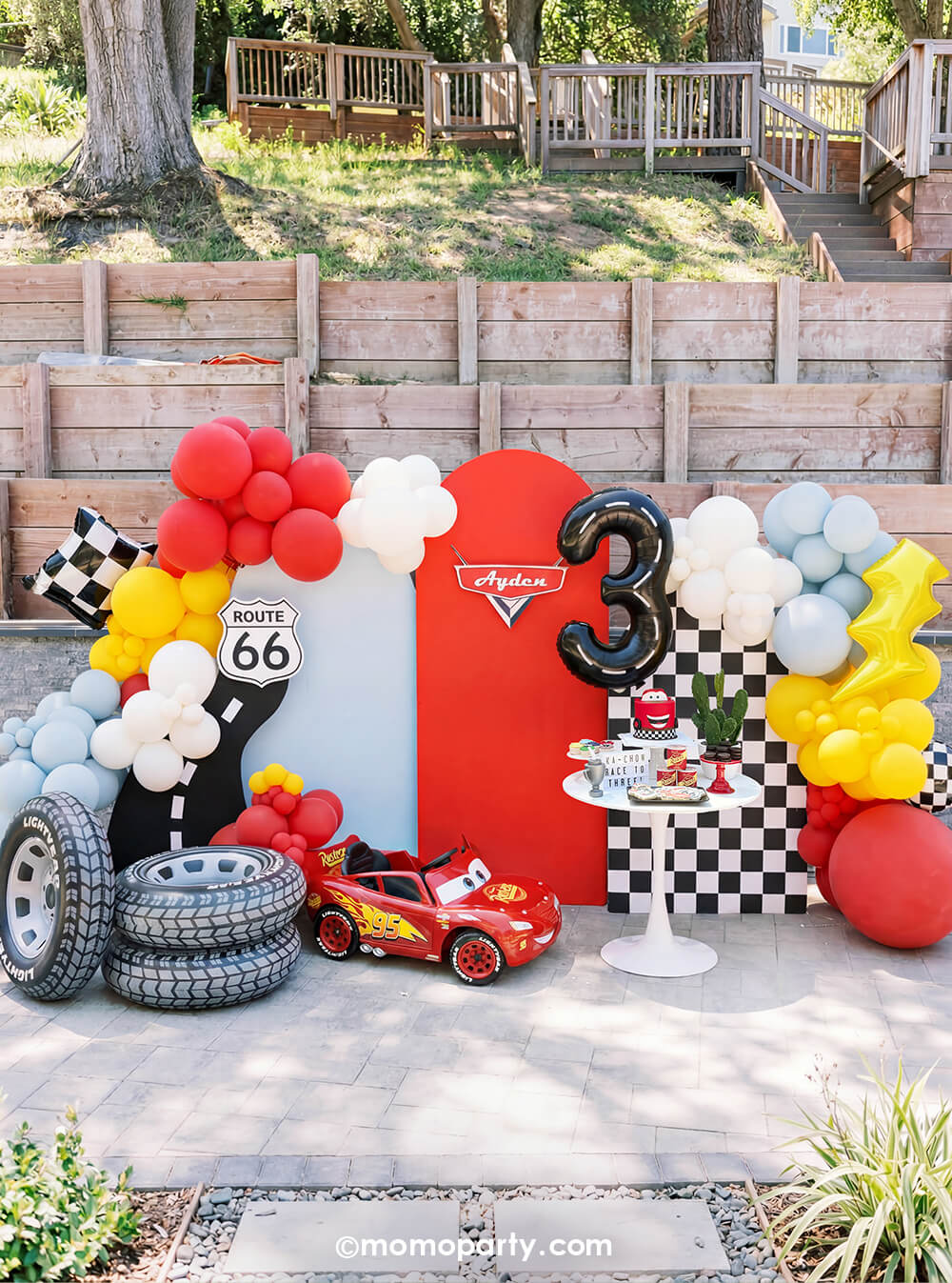 A kid's Disney Cars Lightning Mcqueen themed birthday party set up by Momo Party featuring a table filled with race car themed tableware including race track checkered table runner, fuel treat boxes, red striped cups, checkered plates and mini race car flags as decoration. In teh back is a festive race car themed balloon garland hung around a party backdrop cutout of a race track and Route 66 cutout sign, next to a Lightning Mcqueen race car. A perfect decoration inspo for boy's race car themed birthdays.