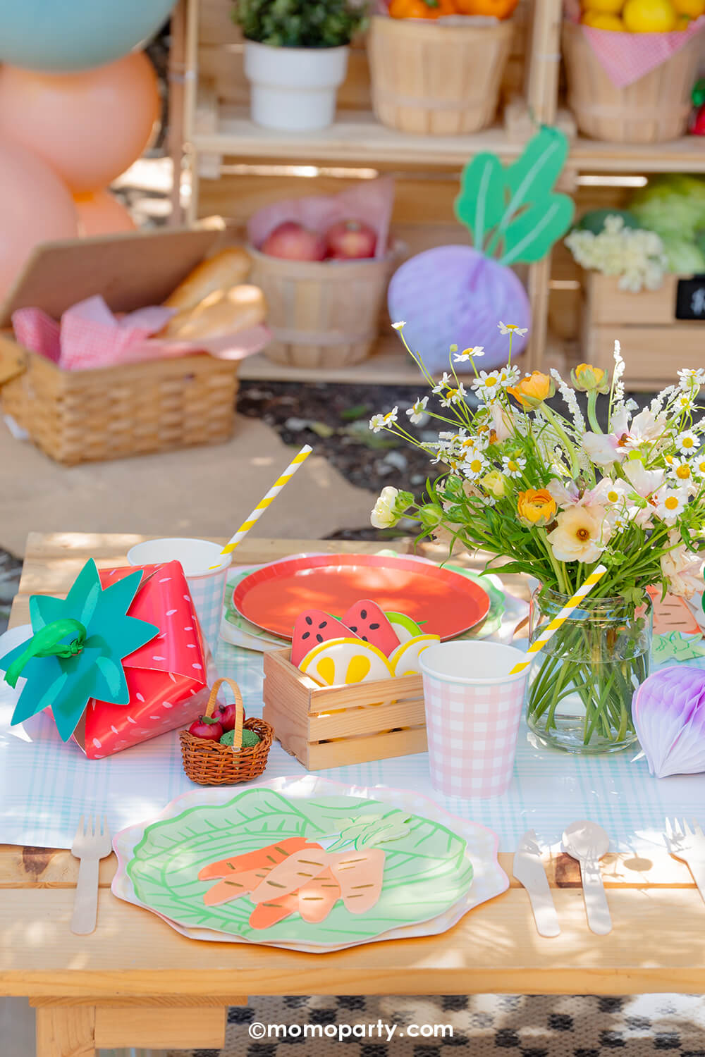 A kid's farmer's market party table setting by Momo Party with cabbage and tomato shaped plates on gingham plate, along with party cups, carrot bunch shaped napkins on a light blue gingham table runner with flowers and a strawberry treat box on a wooden table. On the table there are some sweet treats and in the back is a mini market stall with baskets filled with fruits and veggies, making this a perfect decorating idea for a kid's farmer's market themed birthday, baby shower or a spring/summer gathering.