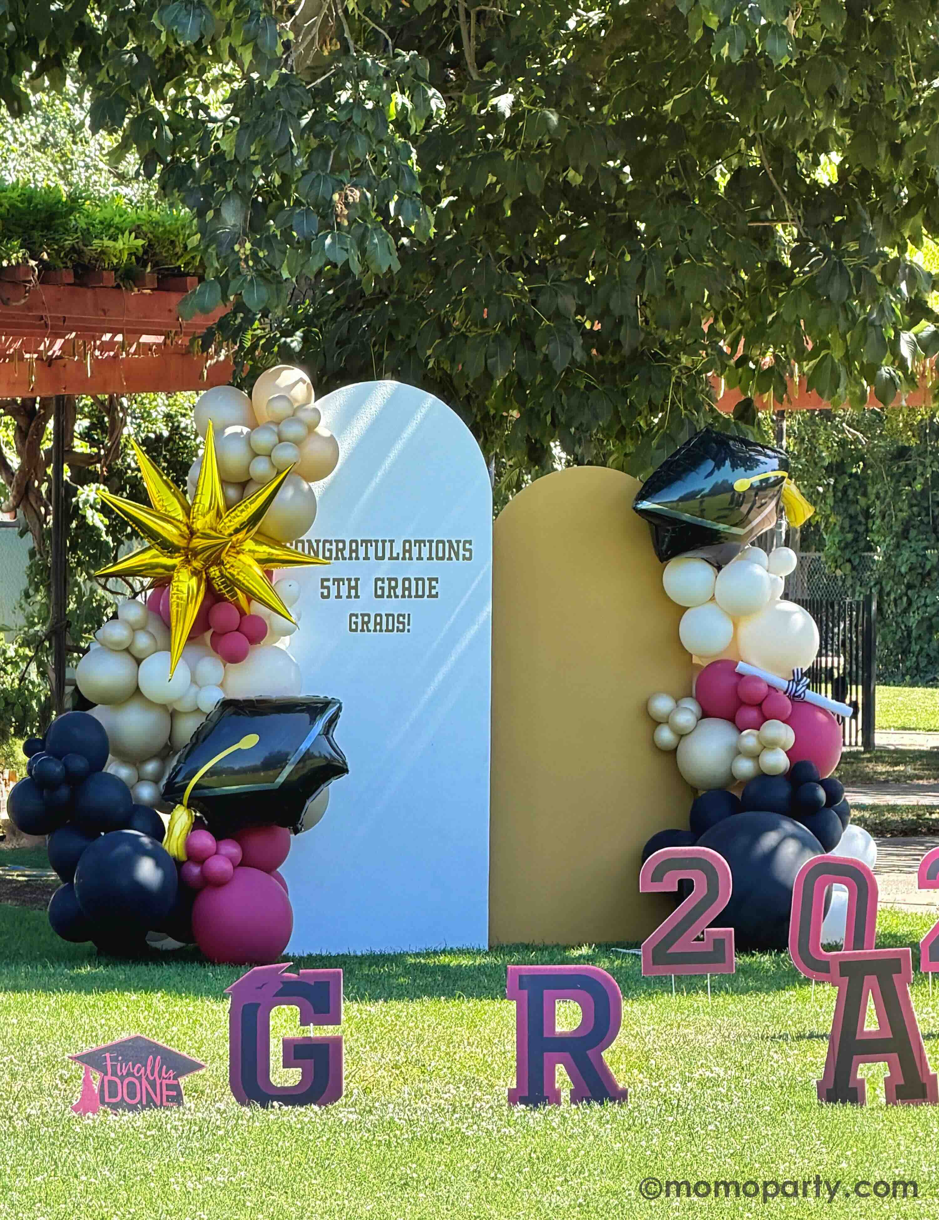 An outdoor elementary school graduation celebration in a park featuring a party backdrop adorn with colorful balloon arch in gold, beige, black and red with two graduation cap foil balloons and a starburst shaped foil balloon next to the 'Congratulations 5th Grade Grads!' sign on the party backdrop, along with 2026 GRAD' letter signs in the front.