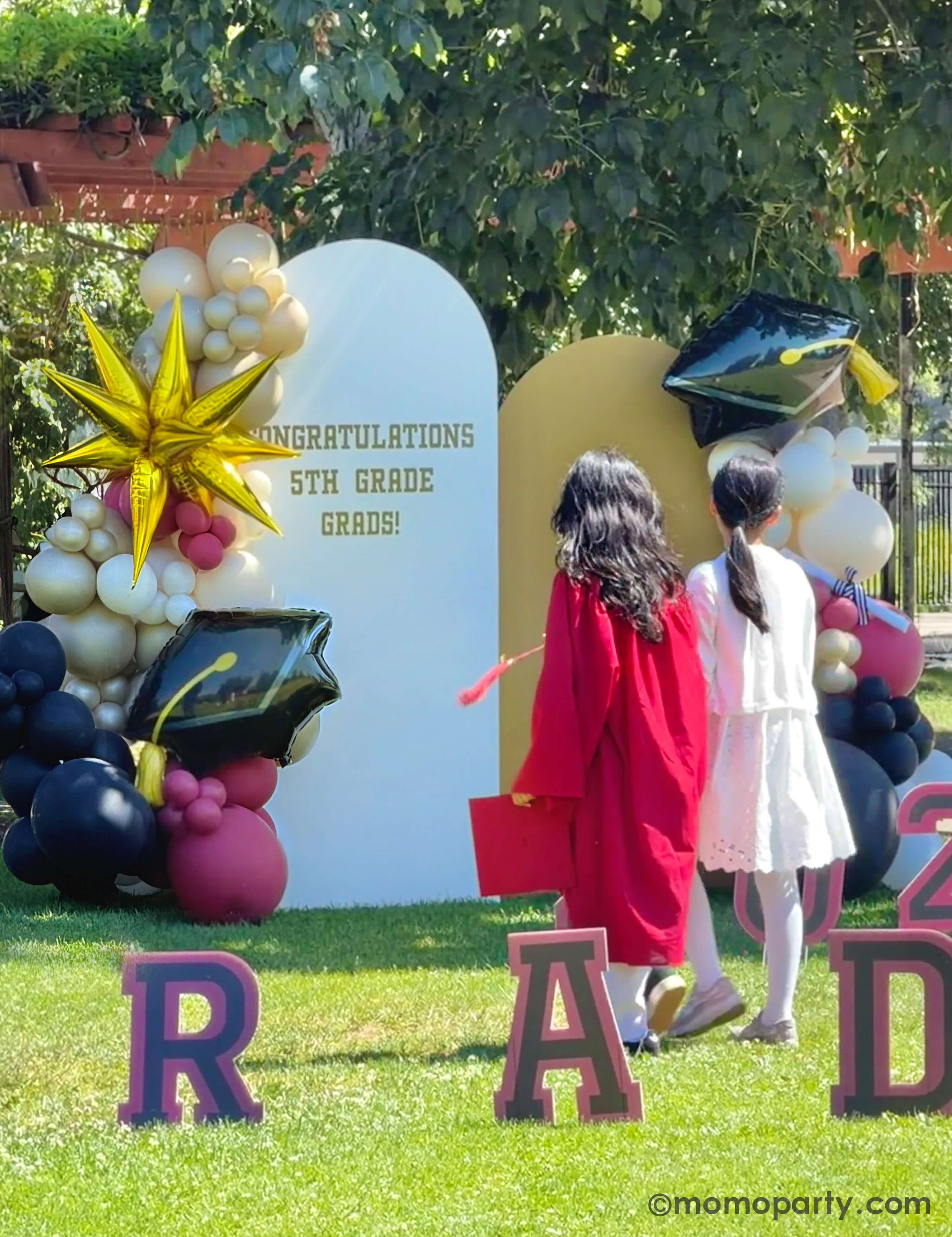 Two children in graduation gowns standing in front of a 'Congratulations 5th Grade Grads!' backdrop with balloons in a graduation celebration in a park featuring a party backdrop adorn with colorful balloon arch in gold, beige, black and red with a graduation cap foil balloon and a starburst shaped foil, along with 'GRAD' letter signs in the front.