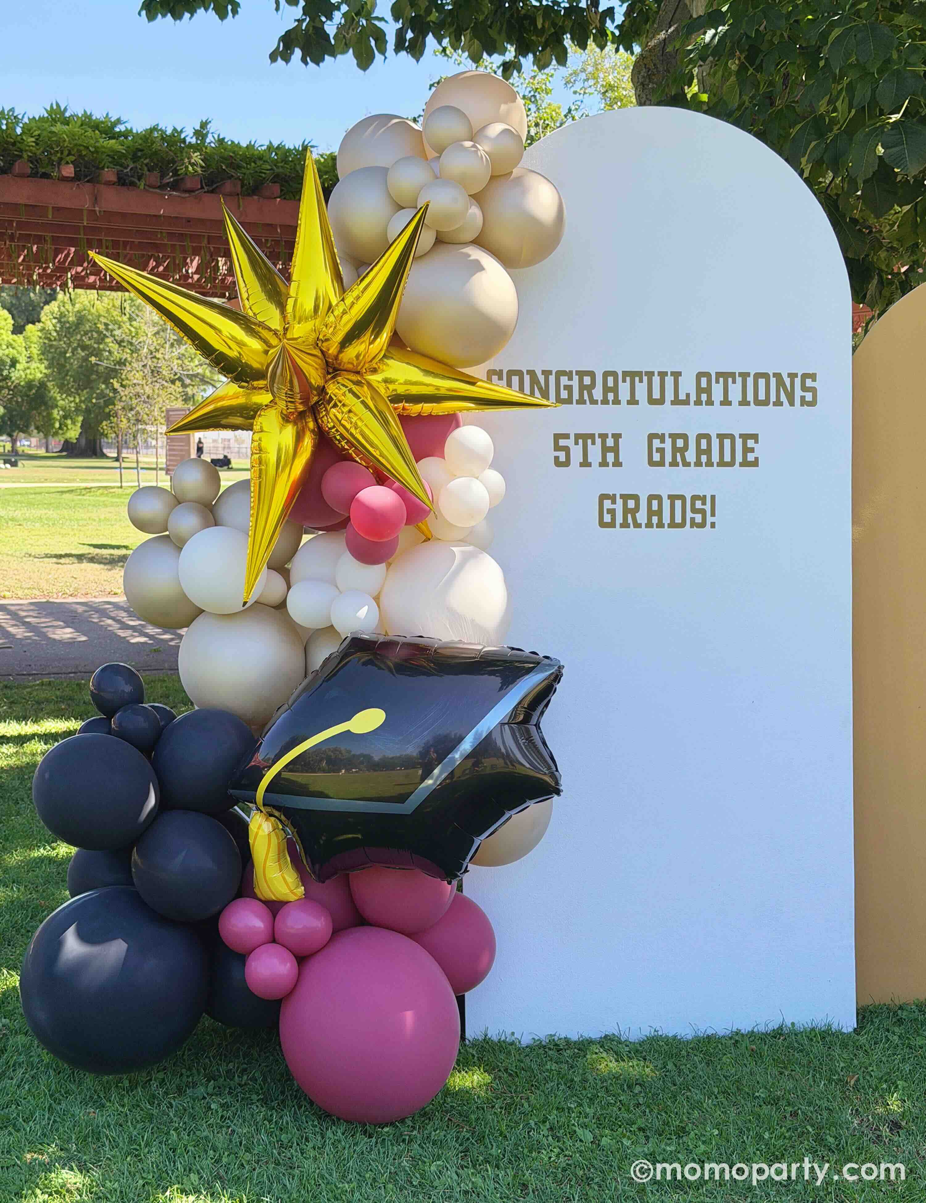 An outdoor elementary school graduation celebration in a park featuring a party backdrop adorn with colorful balloon arch in gold, beige, black and red with a graduation cap foil balloon and a starburst shaped foil balloon next to the 'Congratulations 5th Grade Grads!' sign on the party backdrop.