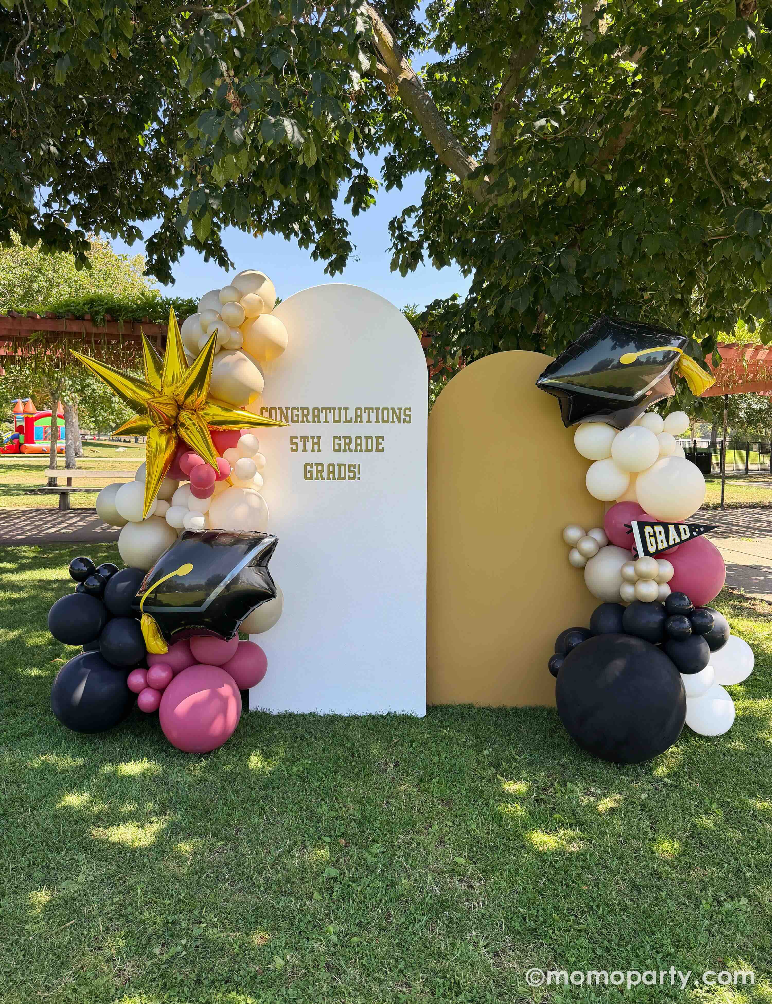 An outdoor elementary school graduation celebration in a park featuring a party backdrop adorn with colorful balloon arch in gold, beige, black and red with two graduation cap foil balloons and a starburst shaped foil balloon next to the 'Congratulations 5th Grade Grads!' sign on the party backdrop on a grassy area.