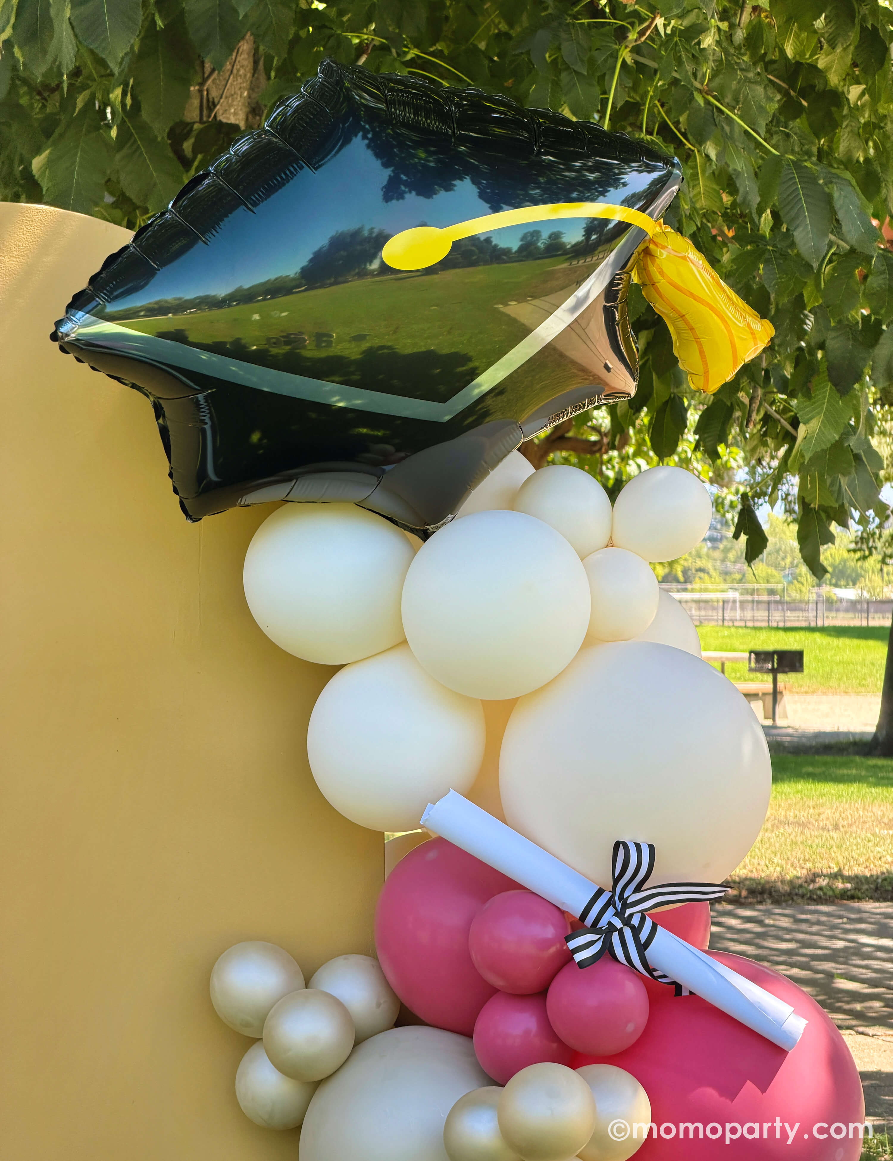 A close up of Momo Party's 31" graduation cap shaped foil balloon in an outdoor elementary school graduation celebration in a park featuring a party backdrop adorn with colorful balloon arch in gold, beige,  and red along with a diploma on the party backdrop.