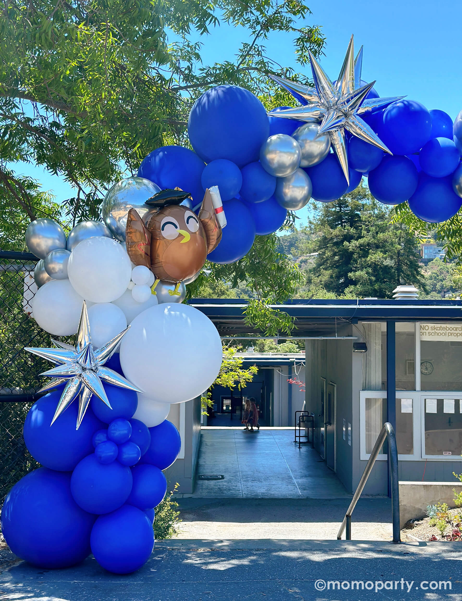 Elementary School Graduation Party by Momo Party. Featuring a stunning balloon arch in school colors of blue, white, and silver, accented with Graduate Owl Shaped Foil Balloons and a 12 Point Silver Starburst Foil Balloon in front of the school staircase. This arch is a big hit, serving as a graduation gate for students to pass through and a photo backdrop to capture last day of school memories.