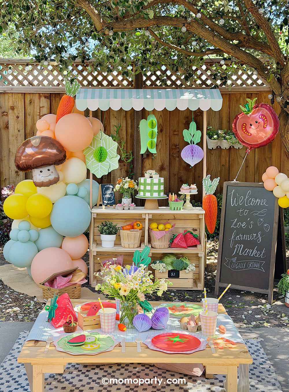 A children's farmer's market themed party outdoor setup by Momo Party with a mini wooden market stall adorned with a colorful pastel balloon garland, produce shaped foil balloons, veggie honeycomb hanging decorations, and food-themed party tablewares on the party table. Along with cakes, cupcakes, sweet treats, floral arrangement, baskets filled with fruits and vegetables, this adorable set up makes a cute kid's birthday party, a baby shower, or a family friendly spring/summer celebration.