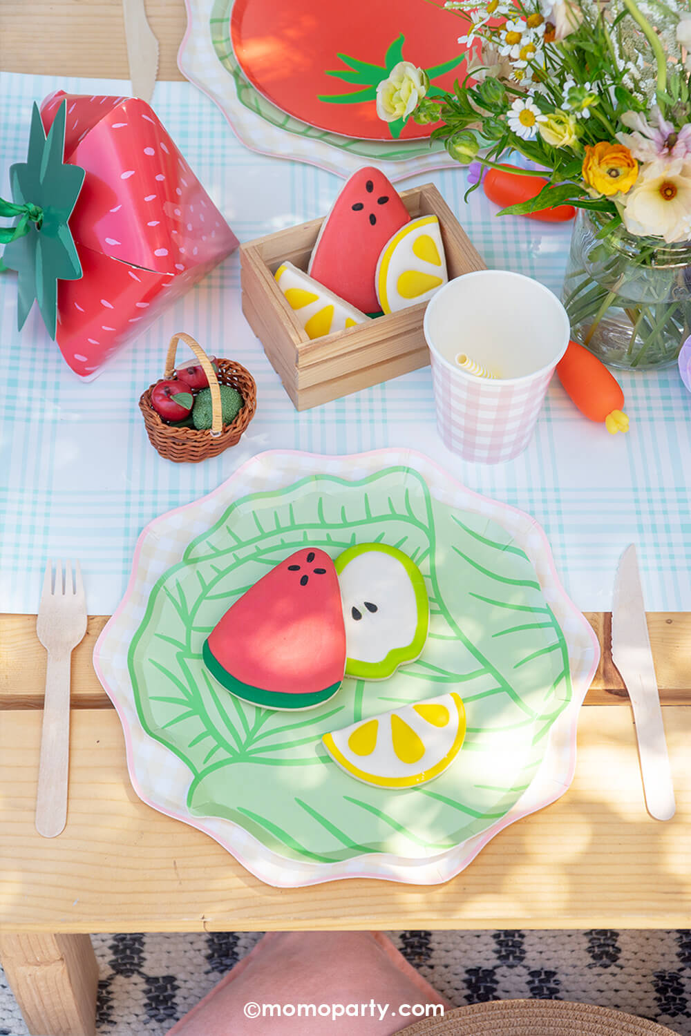 A kid's farmer's market party table setting by Momo Party with cabbage and tomato shaped plates on gingham plates, along with party cups, carrot bunch shaped napkins on a light blue gingham table runner with flowers and a strawberry treat box on a wooden table. On the plates there are some fruit shaped sugar cookies, making this a charming tablescape for a kid's farmer's market themed birthday, baby shower or a spring/summer gathering.