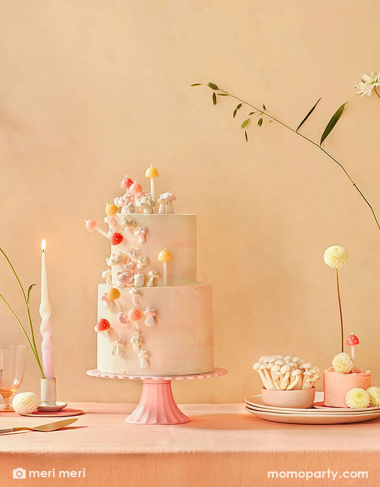 A light pink buttercream cake decorated with Momo Party's 3D mushroom birthday candles by Meri Meri on a pink cake stand. On the table there are also other desserts and some simple spring floral arrangement, a perfect inspo for a modern spring tablescape.