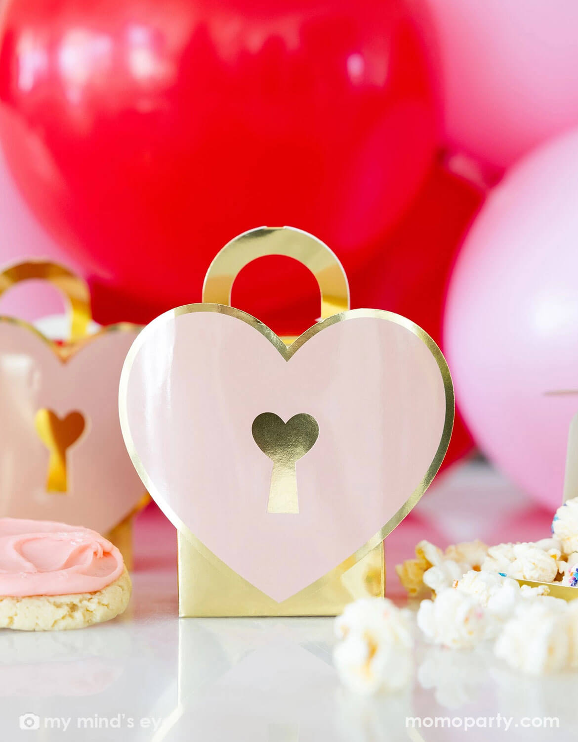 Momo Party's heart-shaped pink and gold favor boxes by My Mind's Eye with red and pink balloons in a Valentine's Day party set up in the background.