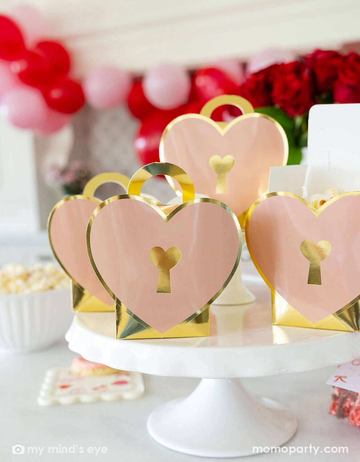 Momo Party's heart-shaped treat boxes with gold accents on a cake stand, surrounded by red and pink balloons and flowers in a festive Valentine's Day party set up.