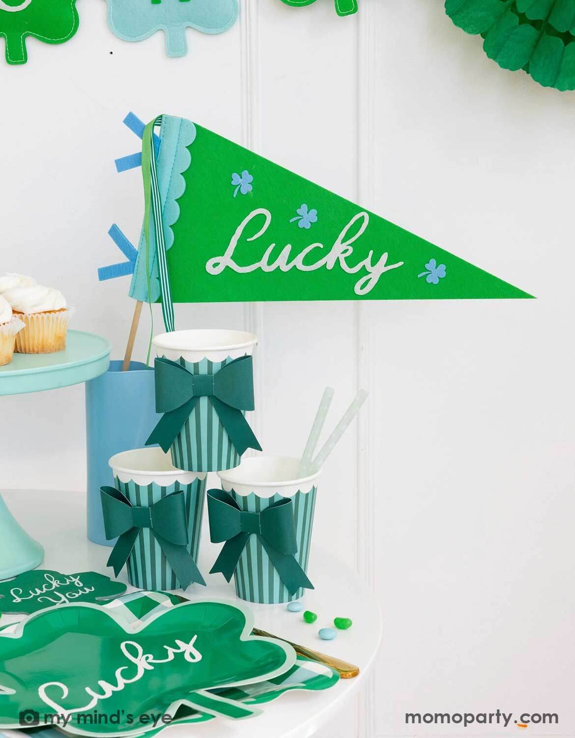 A St. Patrick's Day-themed party set up featuring Momo Party's green 'Lucky' felt party pennant and striped cups with green bows next to a clover shaped plates with 'lucky' text on it.