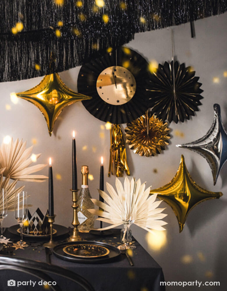 A glamorous black and gold New Year party set up featuring Momo Party's New Year Countdown Party Fans, starpoint shaped balloons and black fringe garlands hung on the wall. In the front is a party table adorned with beautiful black and gold candles, confetti, new year party tableware and floral arrangement, making this a perfect inspiration for a new year eve countdown party.